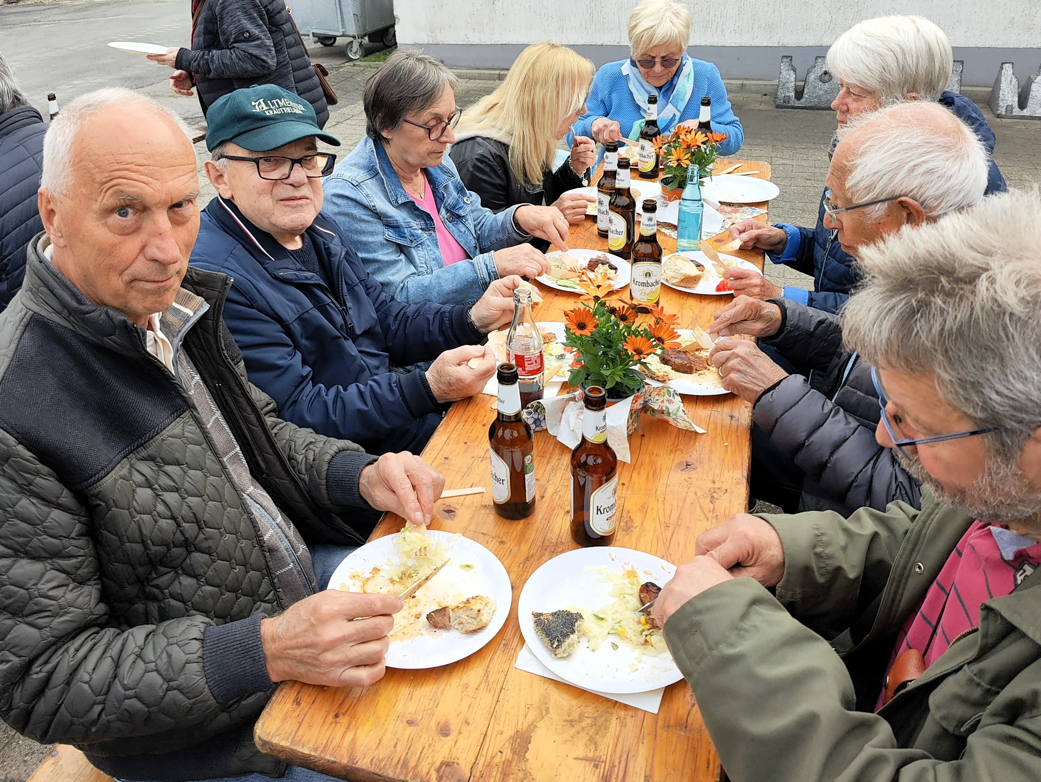 Eine Gruppe älterer Menschen sitzt an einem Holztisch und isst. Auf den Tellern befinden sich Speisen, und es stehen Getränke auf dem Tisch.