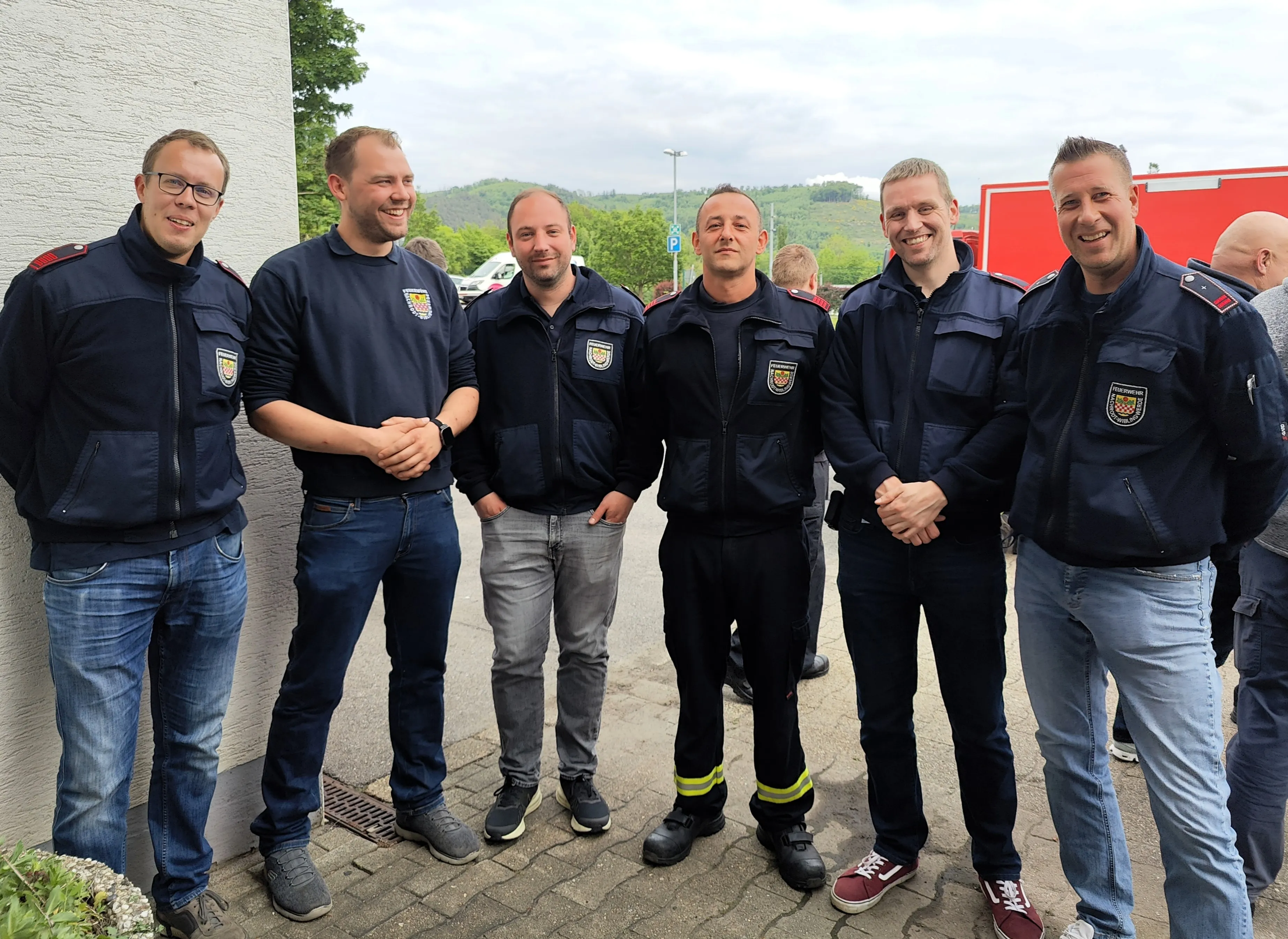 Sechs Feuerwehrleute in Uniform stehen lächelnd vor einer Feuerwache. Im Hintergrund sind weitere Personen und ein bewaldeter Hügel zu sehen.