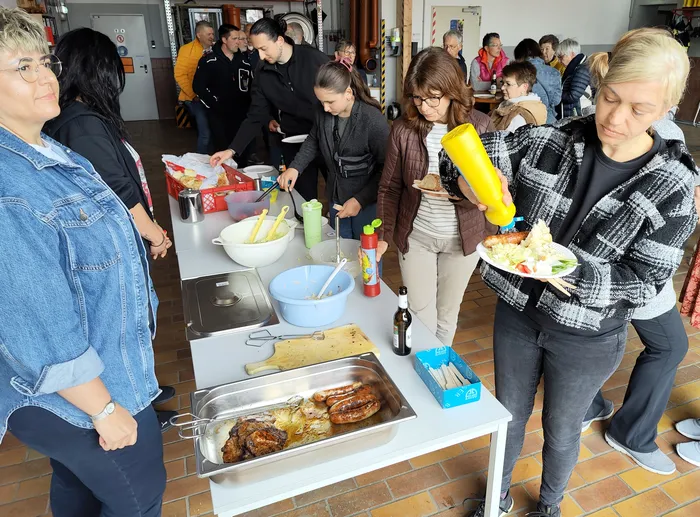 Menschen bedienen sich an einem Grillbuffet mit Würstchen und Salaten bei einer Veranstaltung im Freien.