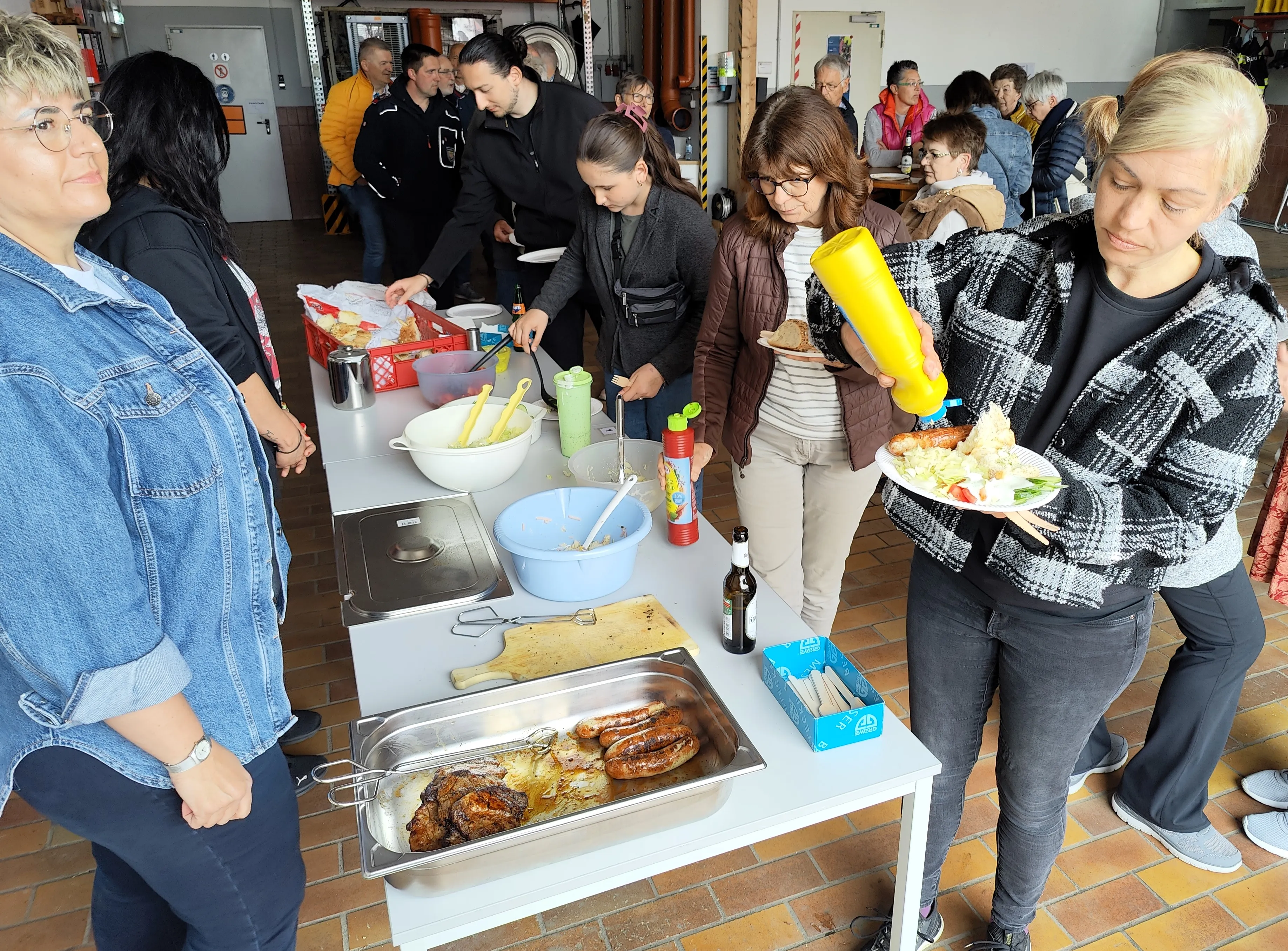 Menschen bedienen sich an einem Grillbuffet mit Würstchen und Salaten bei einer Veranstaltung im Freien.