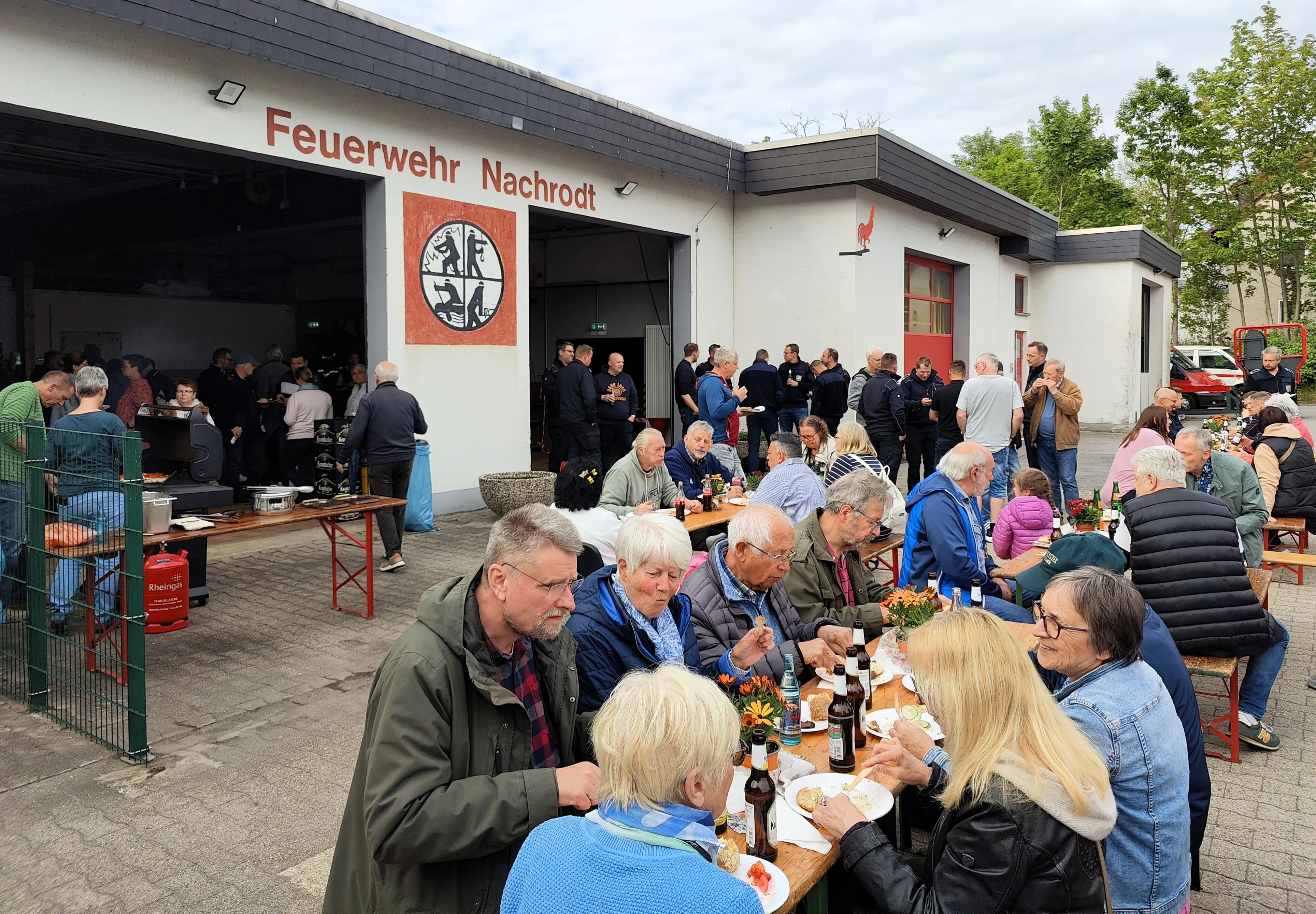 Menschen sitzen an Tischen und essen im Freien vor einem Feuerwehrhaus, während andere im Hintergrund stehen und sich unterhalten.