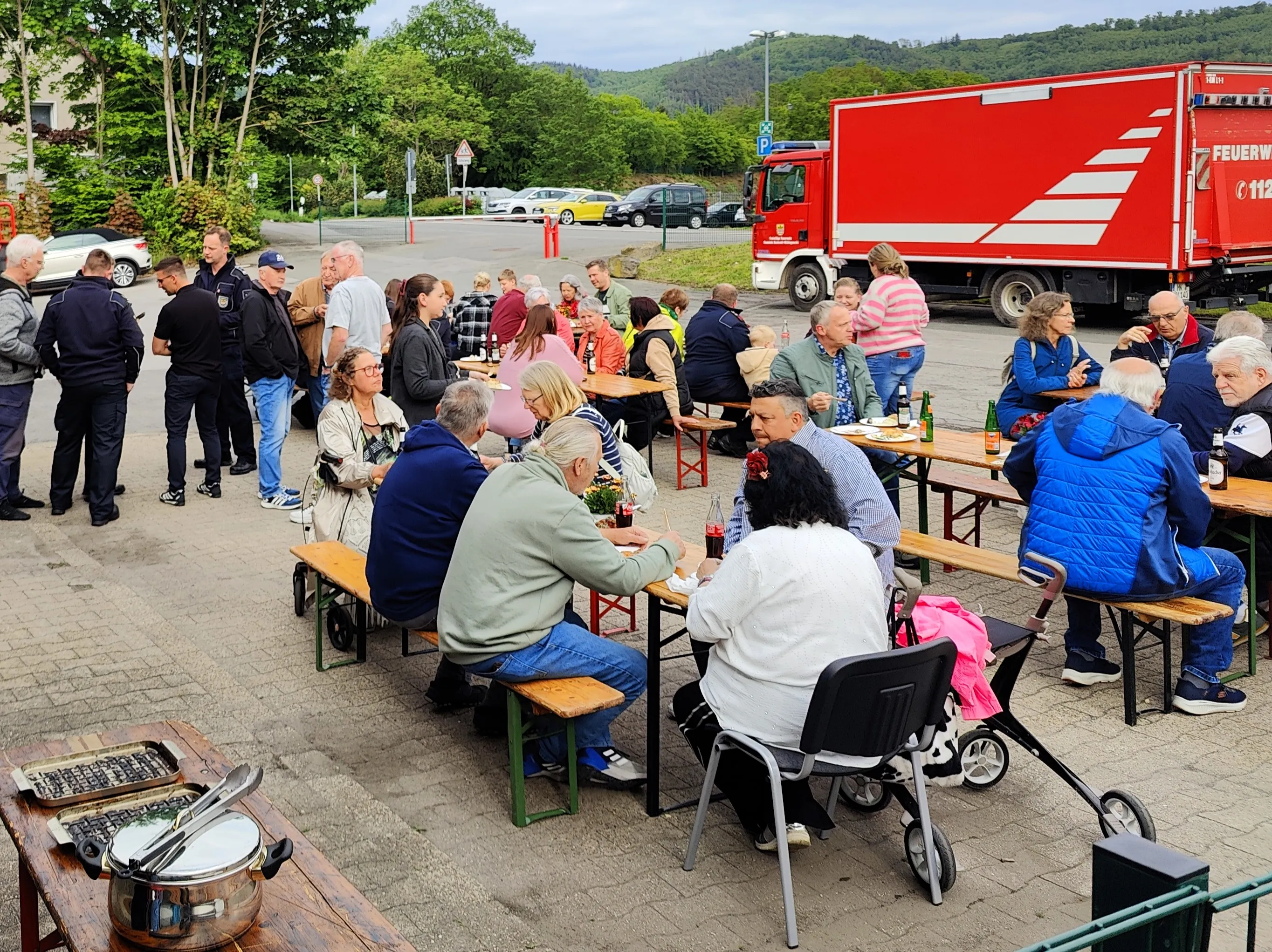 Menschen sitzen und stehen an Biertischen und essen, während im Hintergrund ein rotes Feuerwehrauto steht. Im Vordergrund sind auf einem Tisch Speisen und Getränke arrangiert.