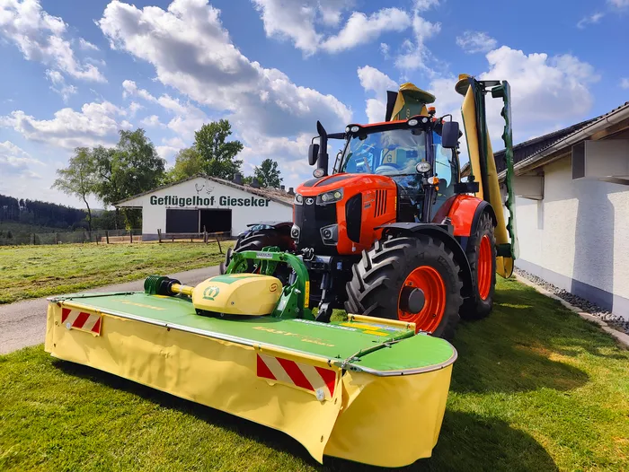 Ein roter Traktor mit einem angebauten Mähwerk steht auf einer grünen Wiese vor einem landwirtschaftlichen Gebäude mit dem Schriftzug „Geflügelhof Gieseker“.
