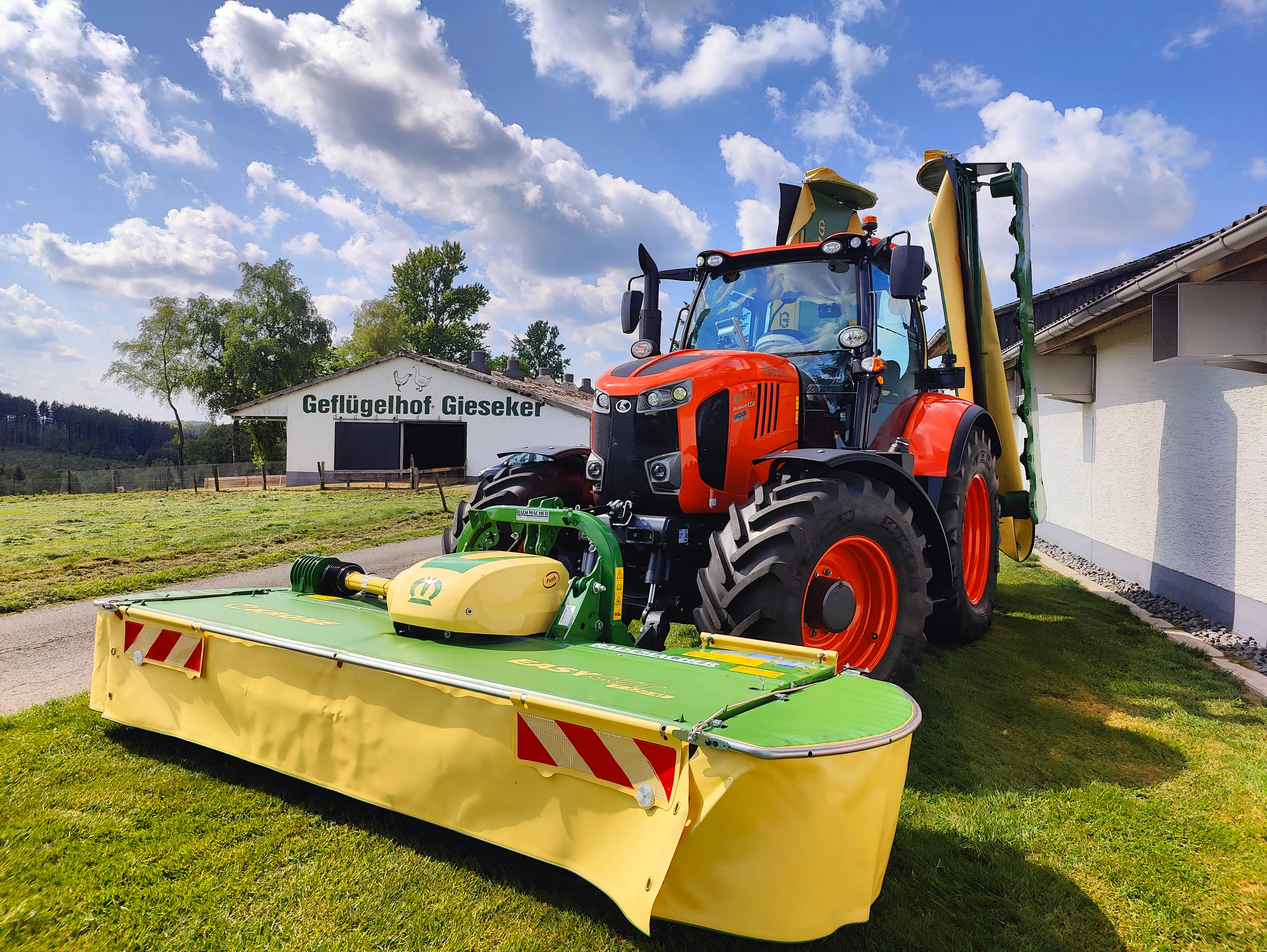 Ein roter Traktor mit einem angebauten Mähwerk steht auf einer grünen Wiese vor einem landwirtschaftlichen Gebäude mit dem Schriftzug „Geflügelhof Gieseker“.