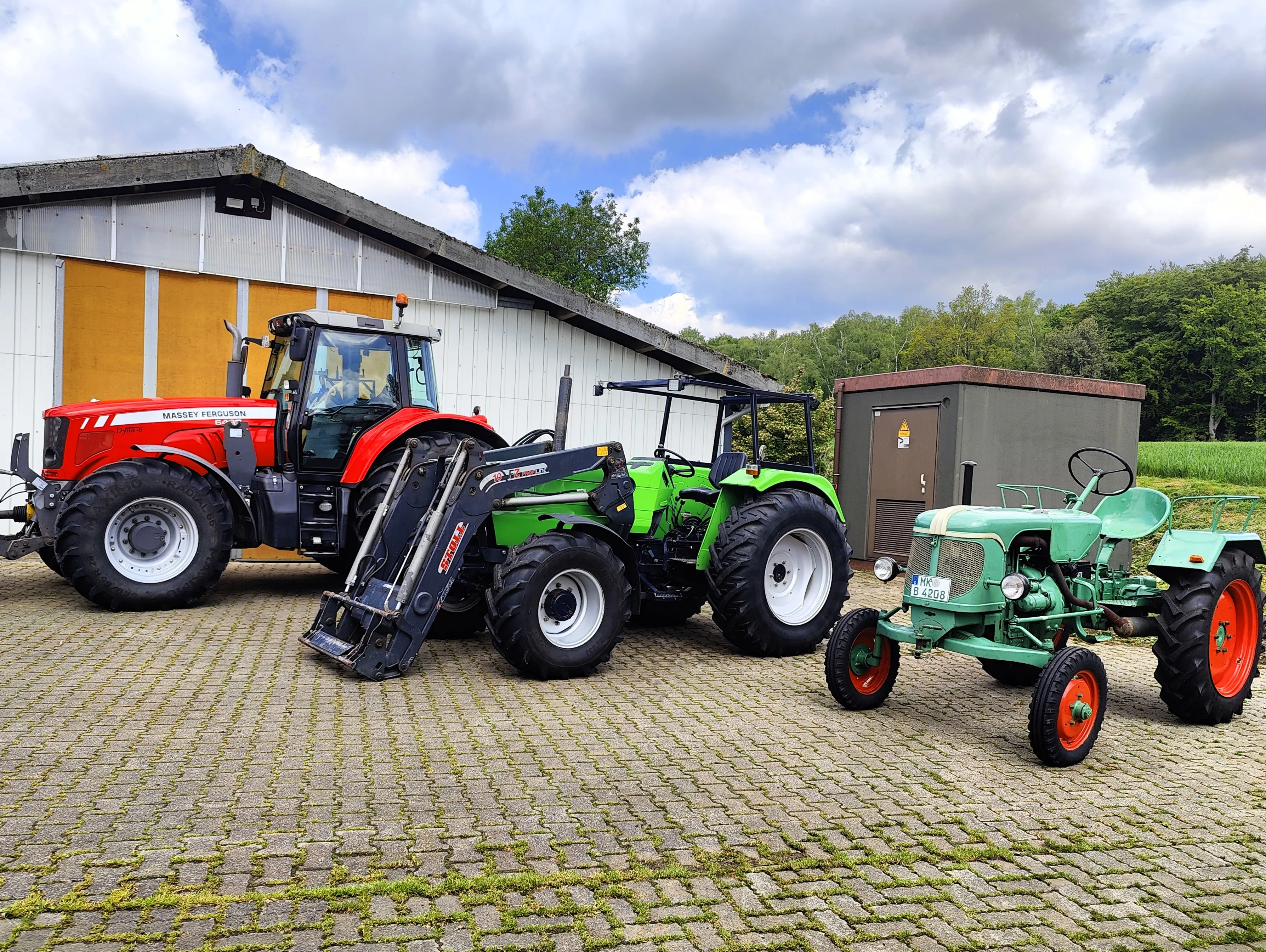 Vier Landwirtschaftsschlepper unterschiedlicher Baujahre stehen auf einem gepflasterten Hof vor einem großen Schuppen.