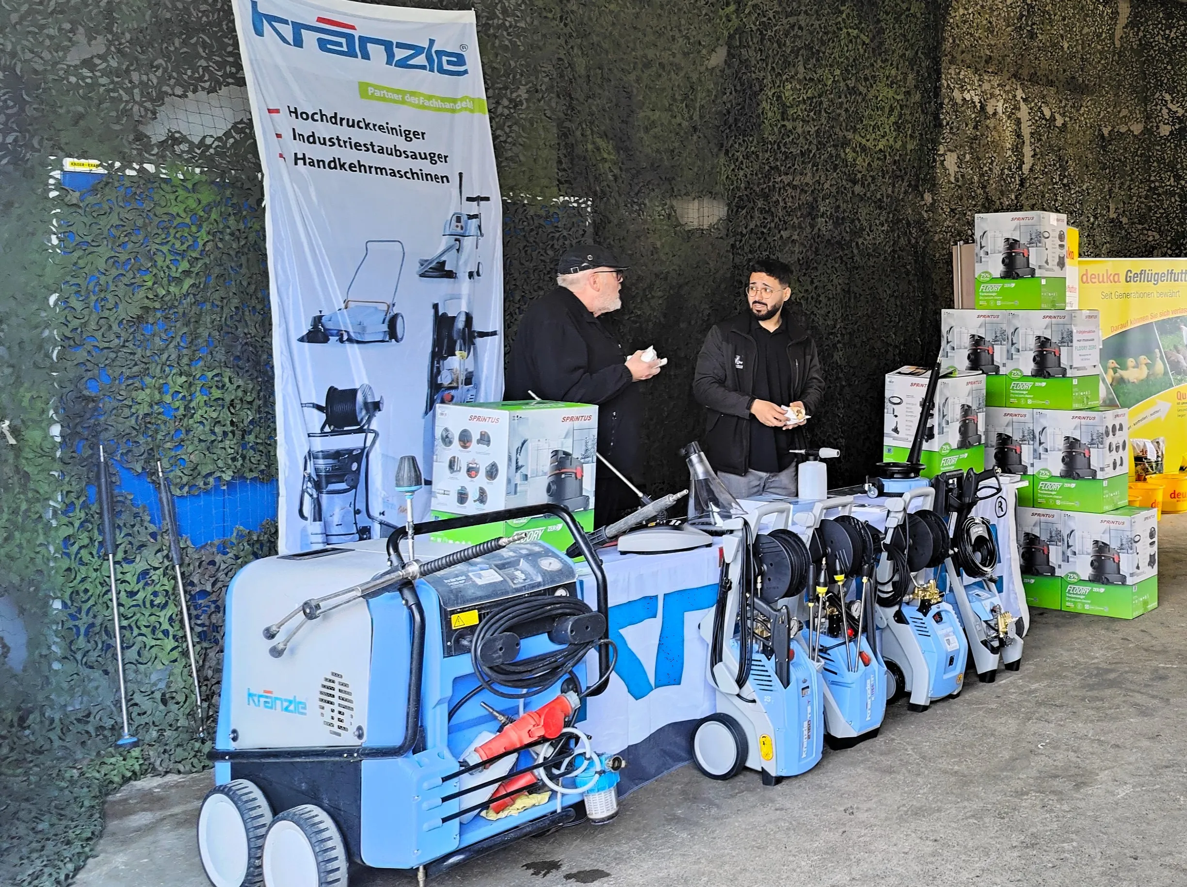 Mehrere blaue Hochdruckreiniger der Marke Kränzle stehen auf einem Messestand vor einer Werbeplane, während ein Mitarbeiter mit einem Kunden spricht.