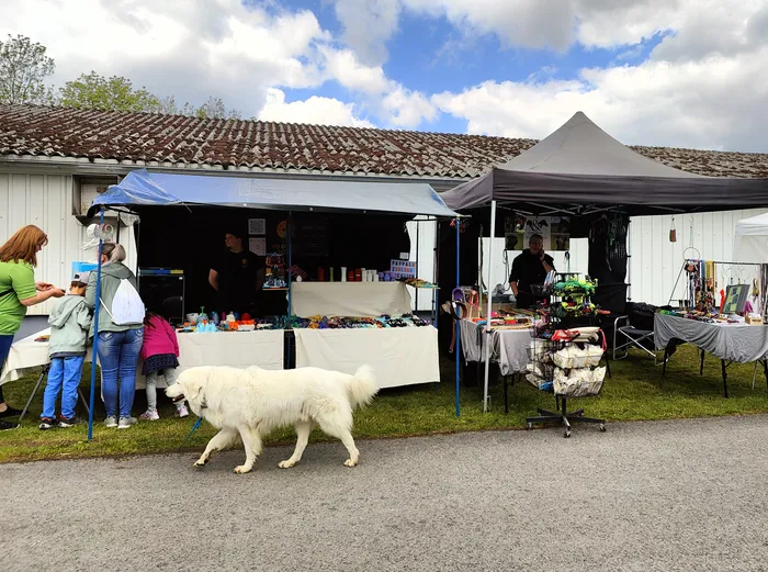 Zwei Verkaufsstände mit bunten Artikeln stehen vor einem Gebäude mit rotem Ziegeldach. Vor den Ständen stehen Menschen und ein großer, weißer Hund.