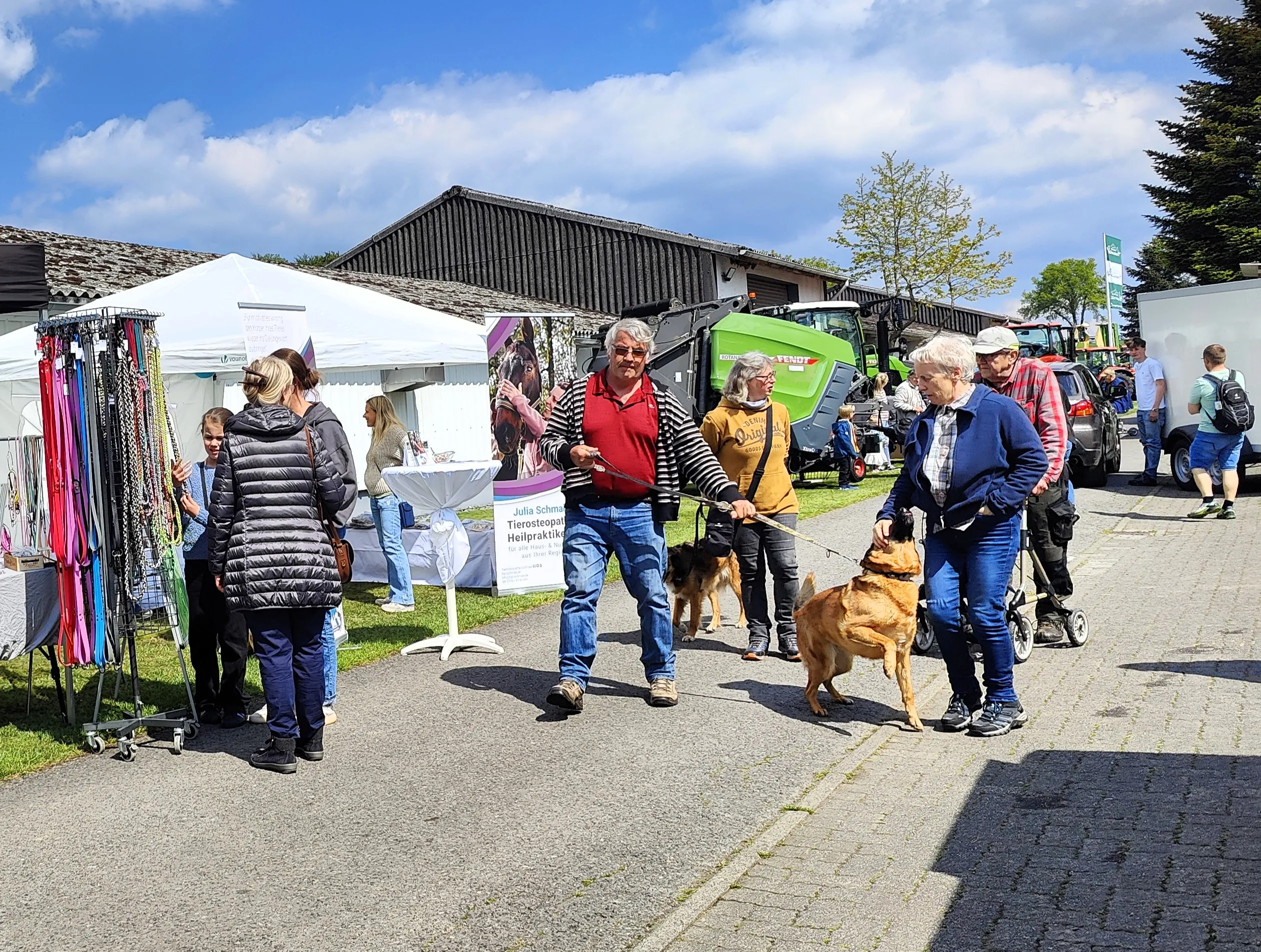 Menschen und ein Hund gehen auf einem Hofgelände an Ständen vorbei, im Hintergrund ein Gebäude und landwirtschaftliche Maschinen.