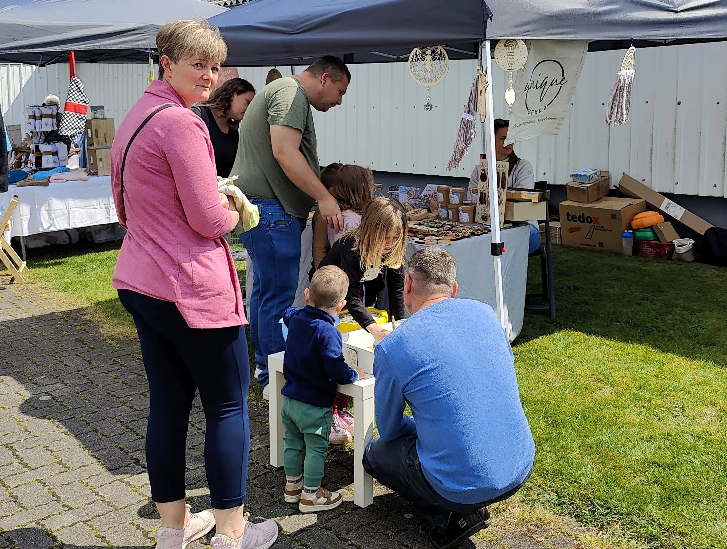 Mehrere Personen betrachten und kaufen Waren an einem Stand auf einem Markt oder einem Bauernhof, darunter auch Kinder.