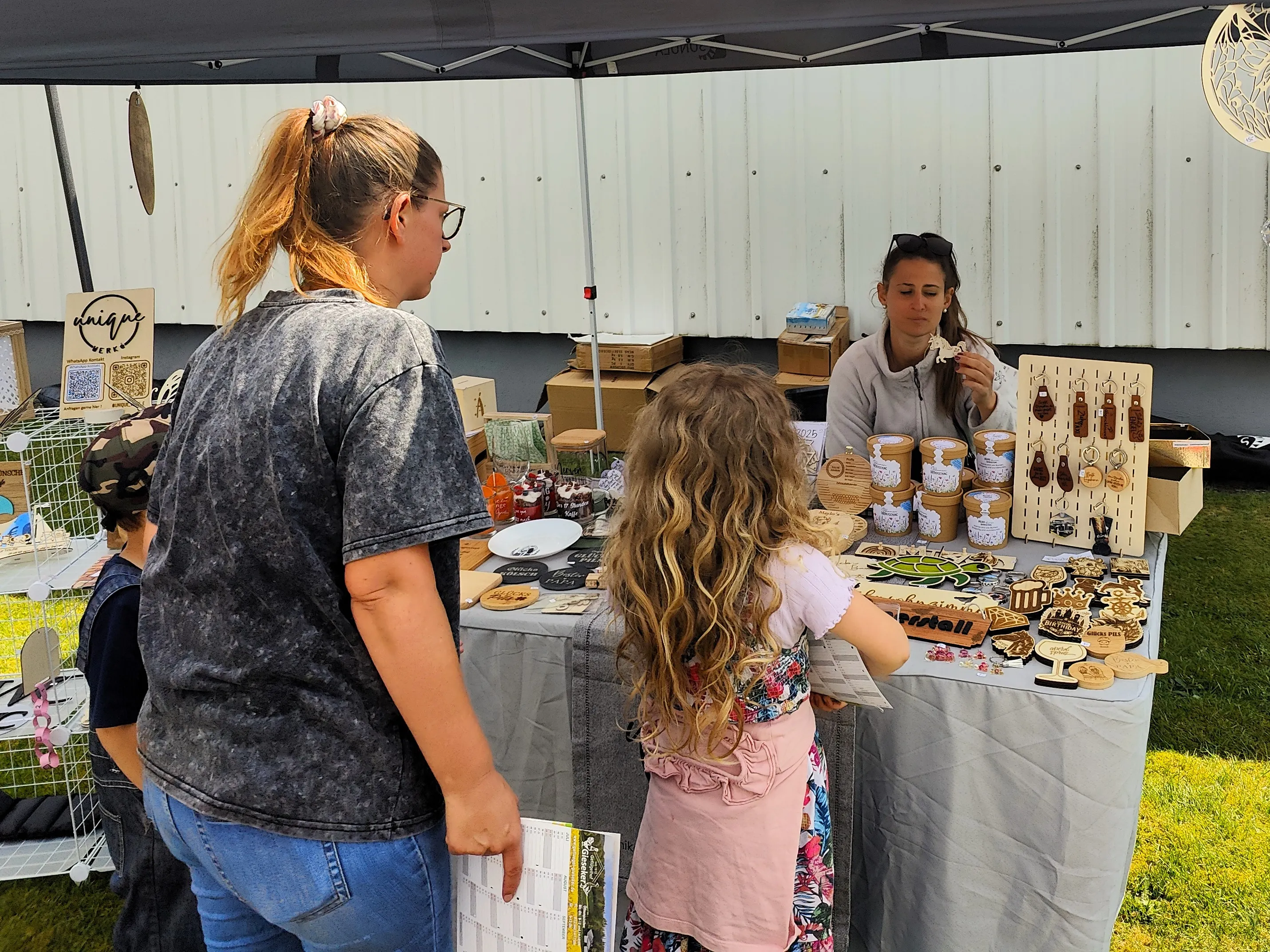 Zwei Frauen und ein Kind betrachten Schmuck und andere Handarbeiten an einem Verkaufsstand auf einem Markt oder Festival.