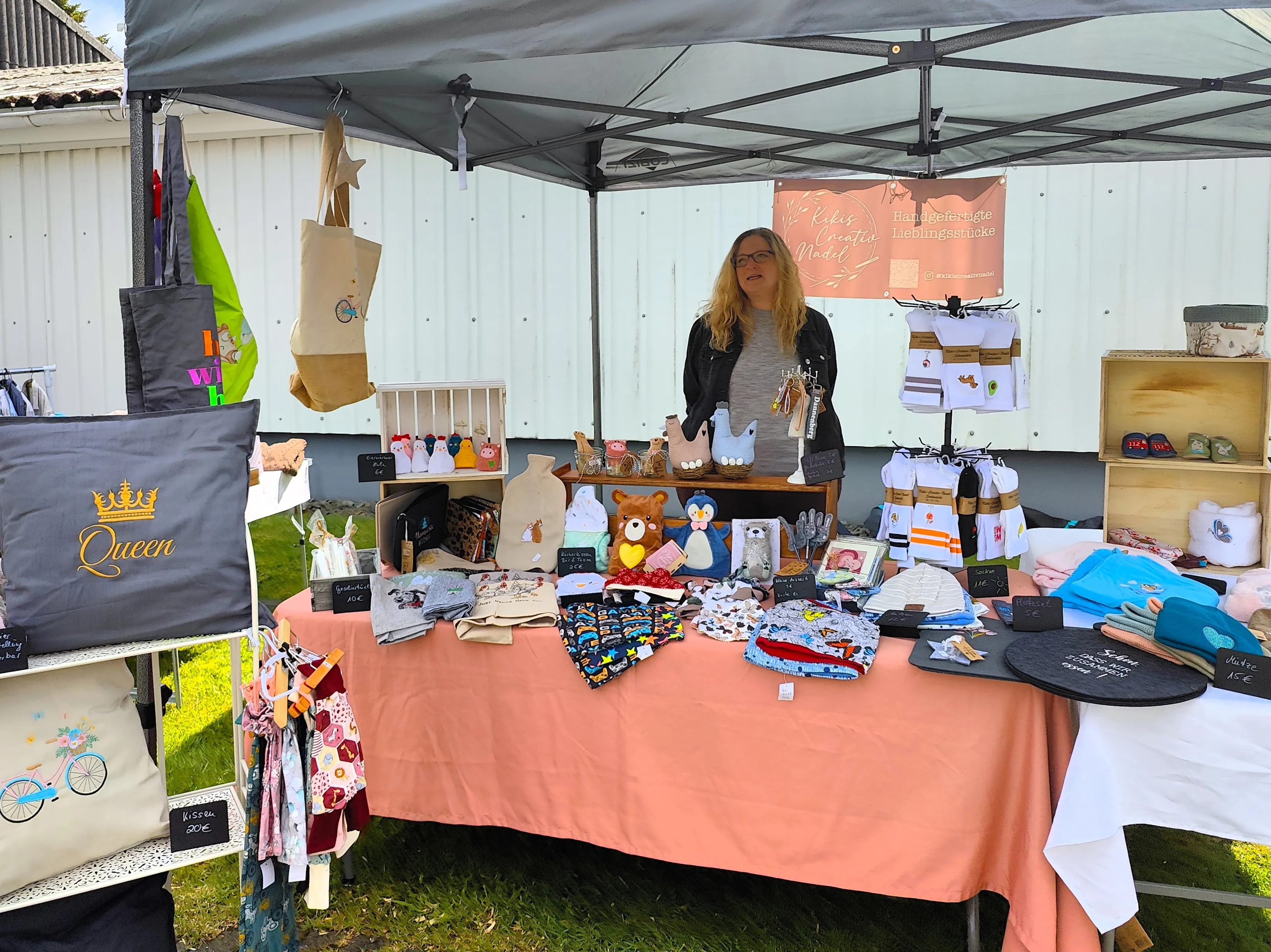Verkäuferin steht an einem Stand mit selbstgemachten Artikeln wie Kissen, Taschen, Stofftieren und Babysachen auf einem Markt im Freien.