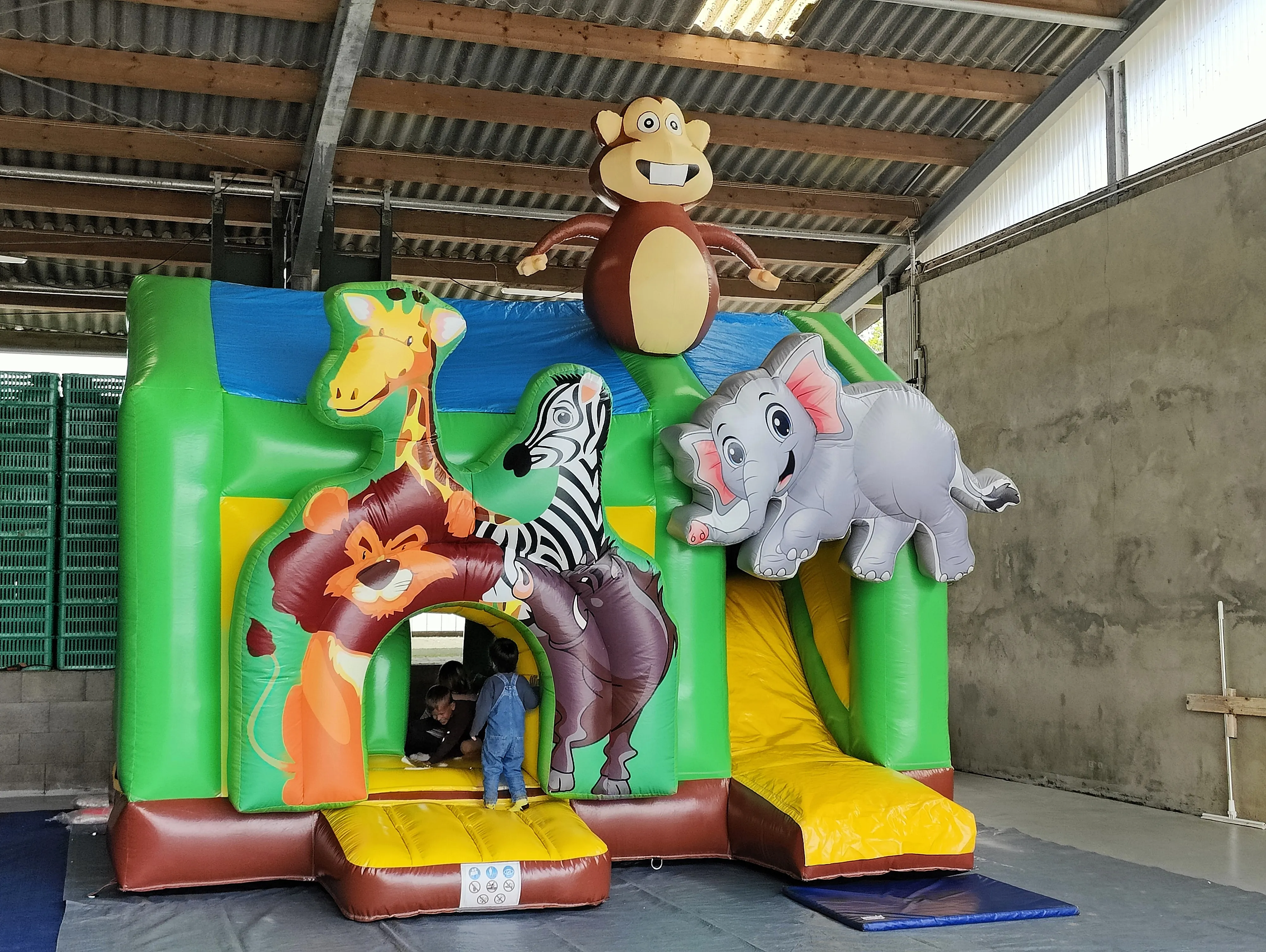 Eine große Hüpfburg mit Tiermotiven steht in einer Lagerhalle. Kinder spielen auf und in der Burg, die einen Affen, einen Elefanten, eine Giraffe, einen Löwen und ein Zebra zeigt.