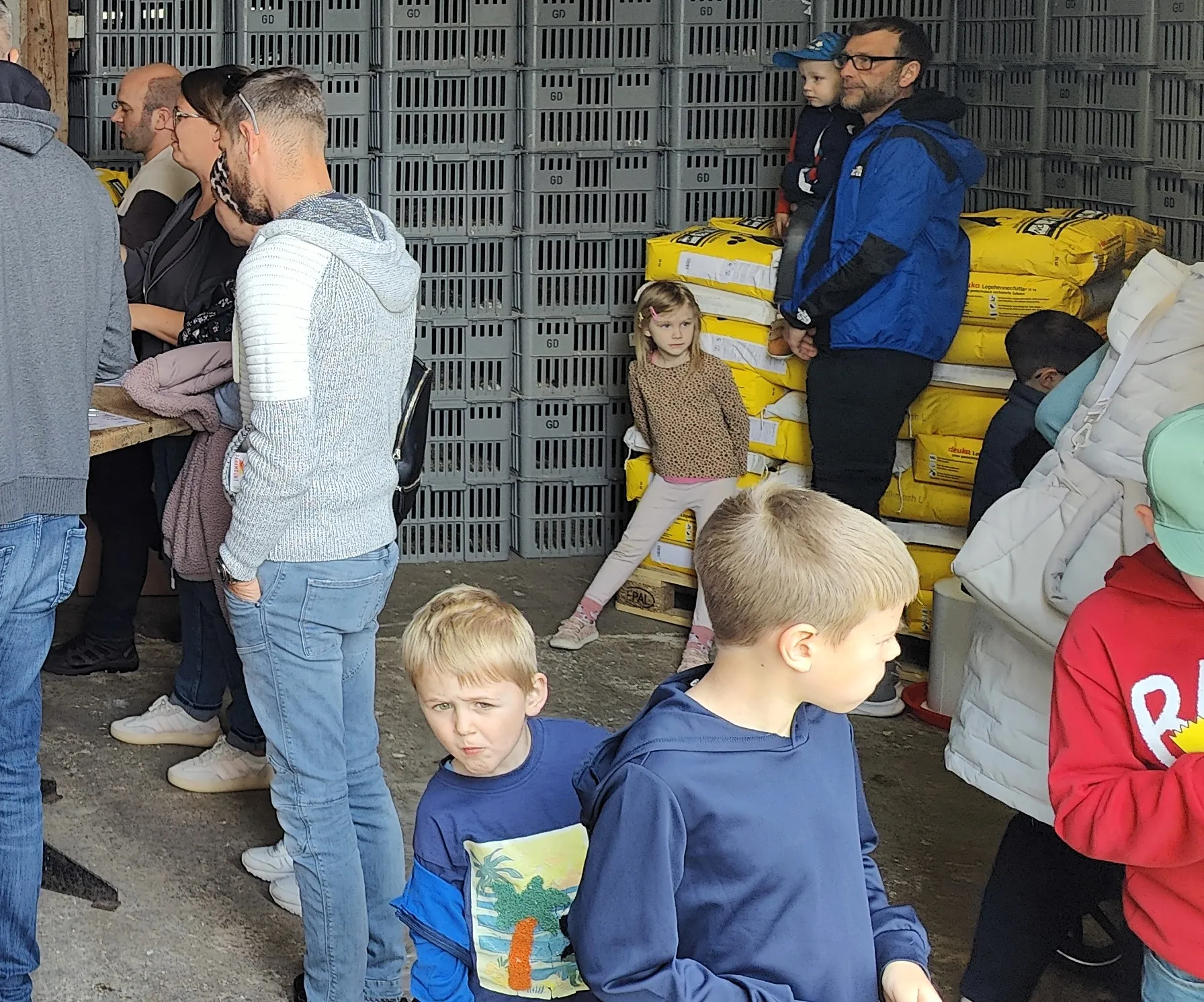 Menschen stehen vor gestapelten Plastikkisten auf dem Gelände eines Geflügelhofs, darunter auch Kinder. Im Hintergrund sind weitere Personen und Kisten zu sehen.