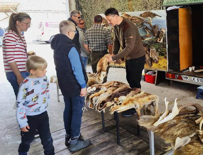 Mehrere Personen betrachten Felle und Schädel auf einem Stand, vor dem ein Mann in brauner Jacke steht und etwas erklärt. Im Hintergrund ist ein Anhänger mit einem Bild eines Raubvogels zu sehen.