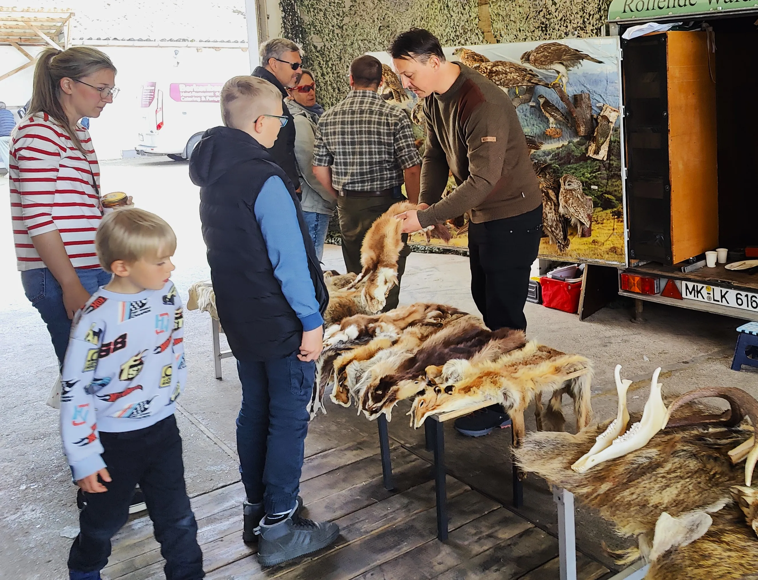 Mehrere Personen betrachten Felle und Schädel auf einem Stand, vor dem ein Mann in brauner Jacke steht und etwas erklärt. Im Hintergrund ist ein Anhänger mit einem Bild eines Raubvogels zu sehen.