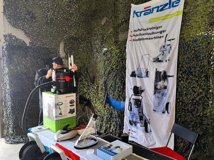 Ein Mann steht hinter einem Tisch mit Reinigungsgeräten und Zubehör auf einem Geflügelhof. Im Hintergrund ein Banner der Marke Kränzle mit Abbildungen von Hochdruckreinigern und Staubsaugern.