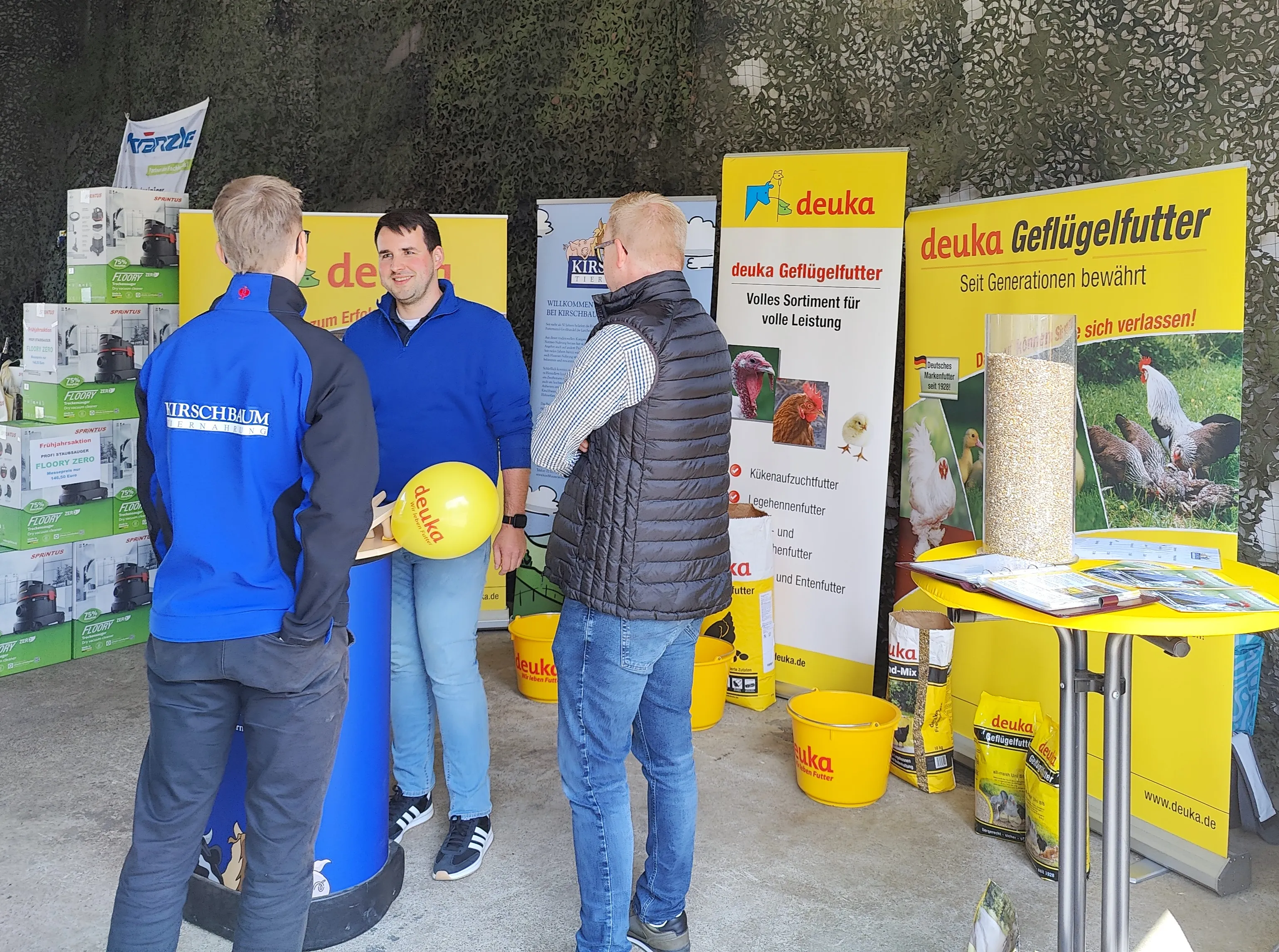 Drei Personen betrachten an einem Stand mit Geflügelfutterprodukten der Marke deuka verschiedene Produkte und lassen sich beraten.