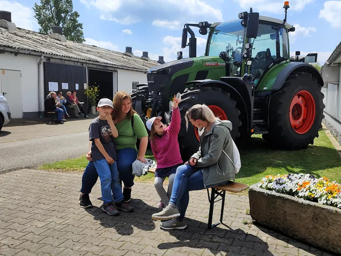 Drei Frauen und ein Kind sitzen vor einem großen grünen Traktor und schauen nach oben. Im Hintergrund sind weitere Menschen und Gebäude eines Bauernhofs zu sehen.