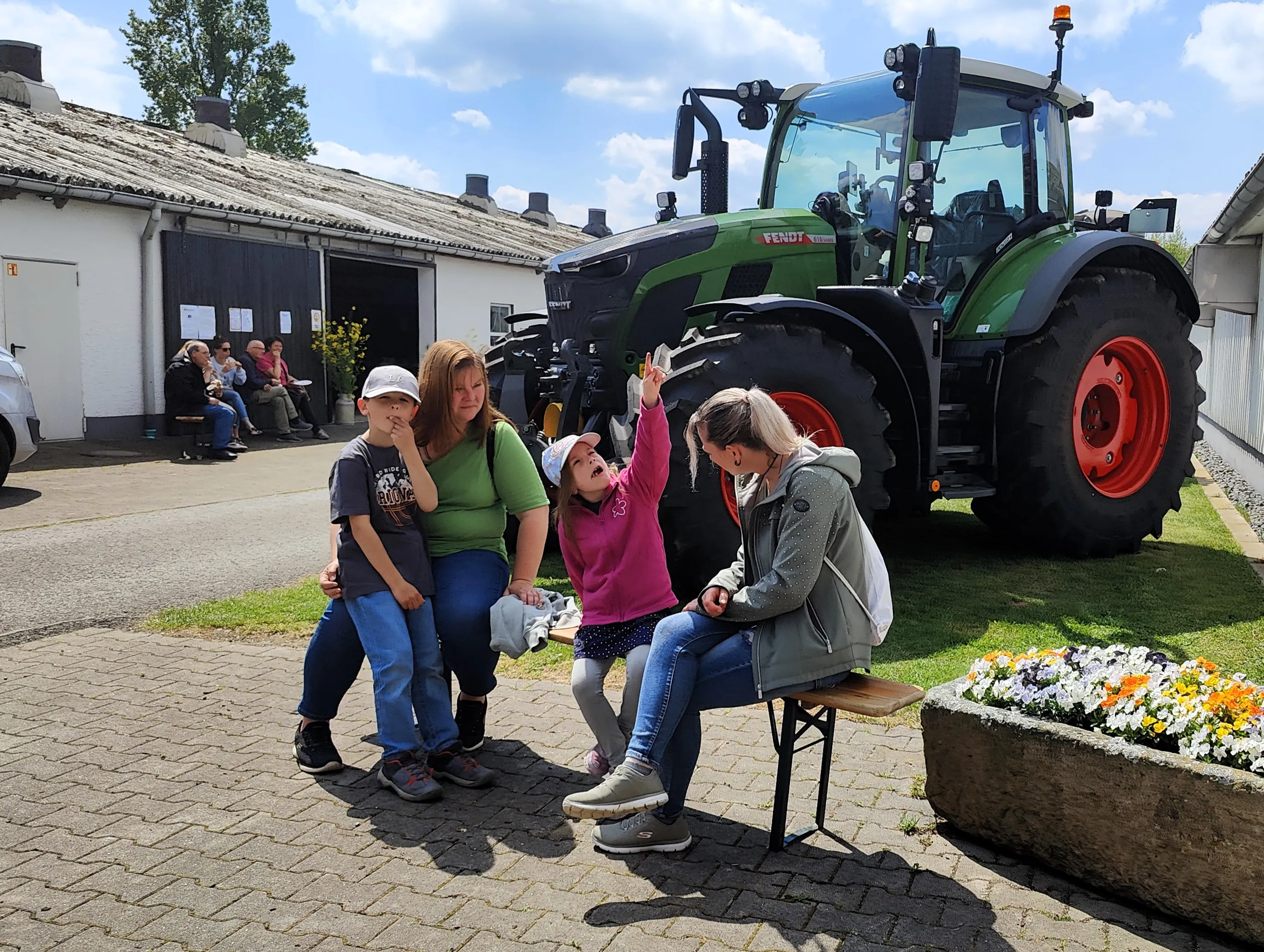 Drei Frauen und ein Kind sitzen vor einem großen grünen Traktor und schauen nach oben. Im Hintergrund sind weitere Menschen und Gebäude eines Bauernhofs zu sehen.