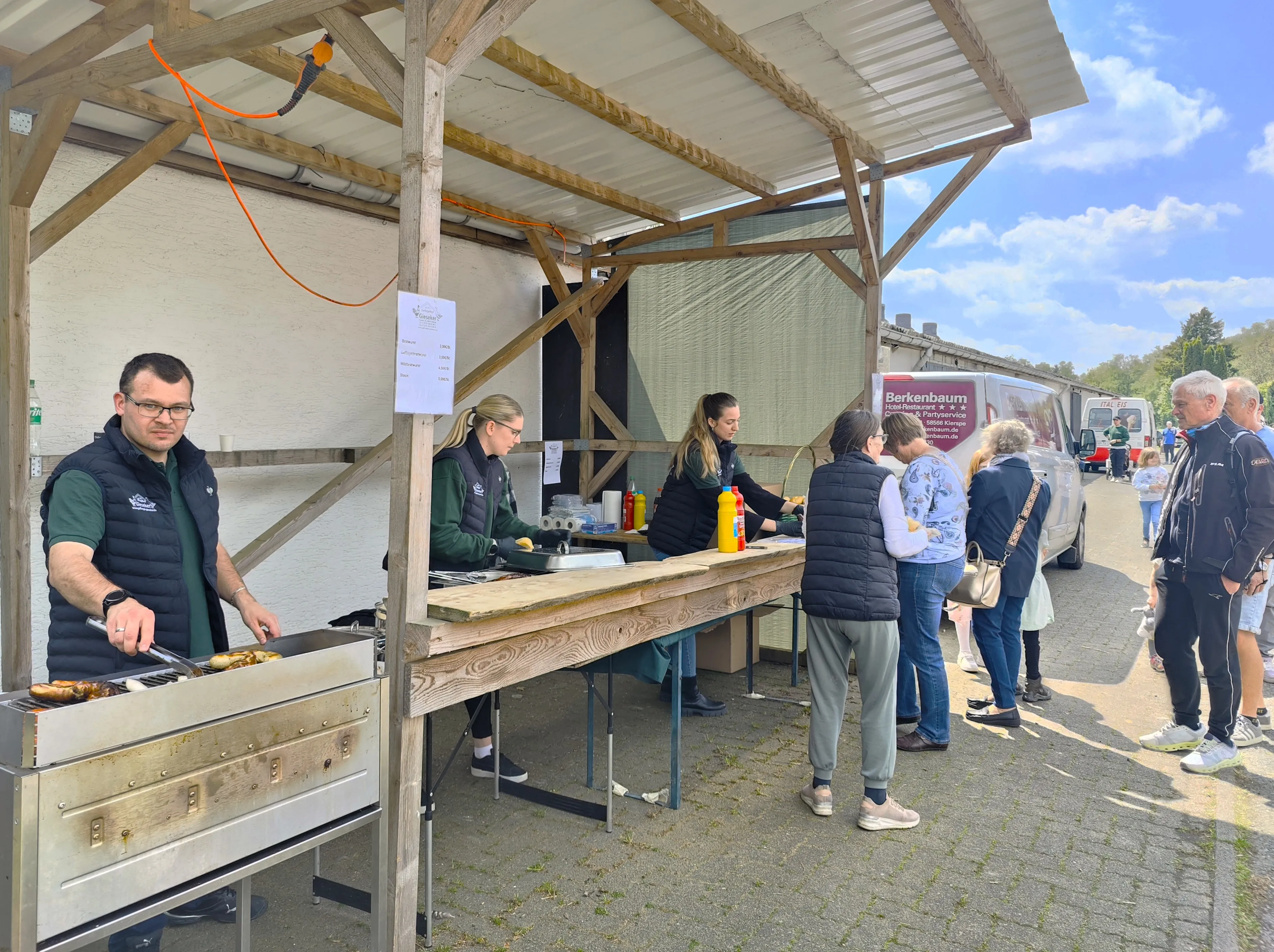 Ein Mann grillt Fleisch auf einem Holzkohlegrill vor einem Imbissstand, während eine Schlange von Menschen auf Essen wartet. Im Hintergrund steht ein Wohnmobil mit der Aufschrift „Berkenbaum“.