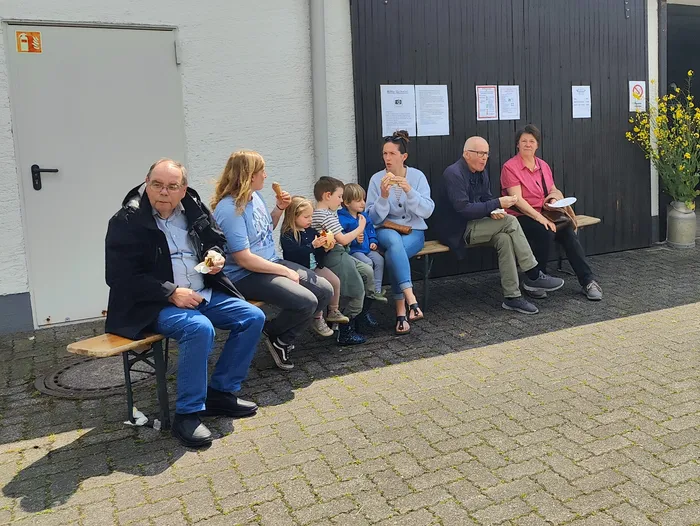 Eine Gruppe von Menschen sitzt auf einer Bank vor einem Gebäude und isst Eis. Im Hintergrund sind Plakate an einer Tür zu sehen.
