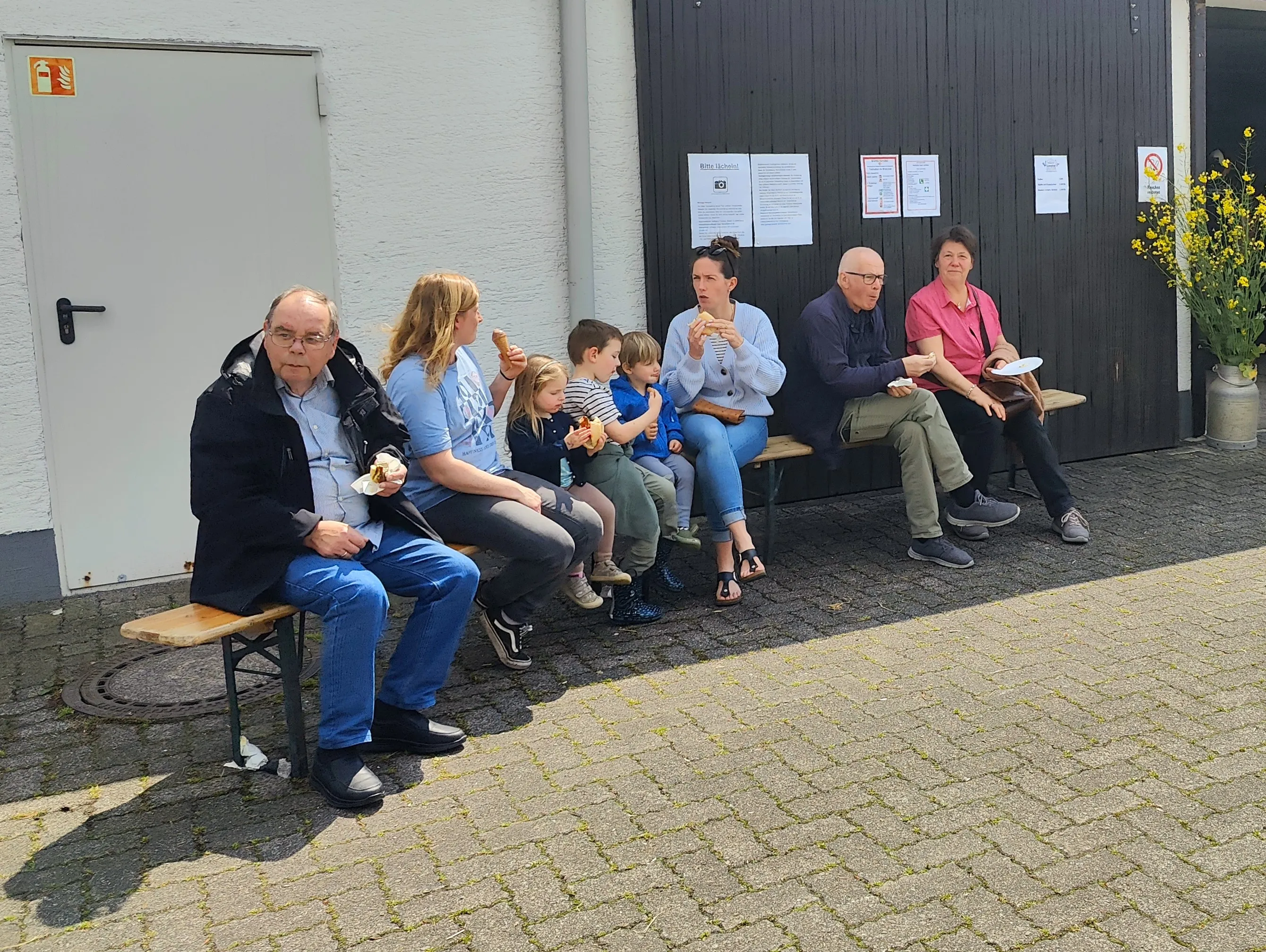 Eine Gruppe von Menschen sitzt auf einer Bank vor einem Gebäude und isst Eis. Im Hintergrund sind Plakate an einer Tür zu sehen.