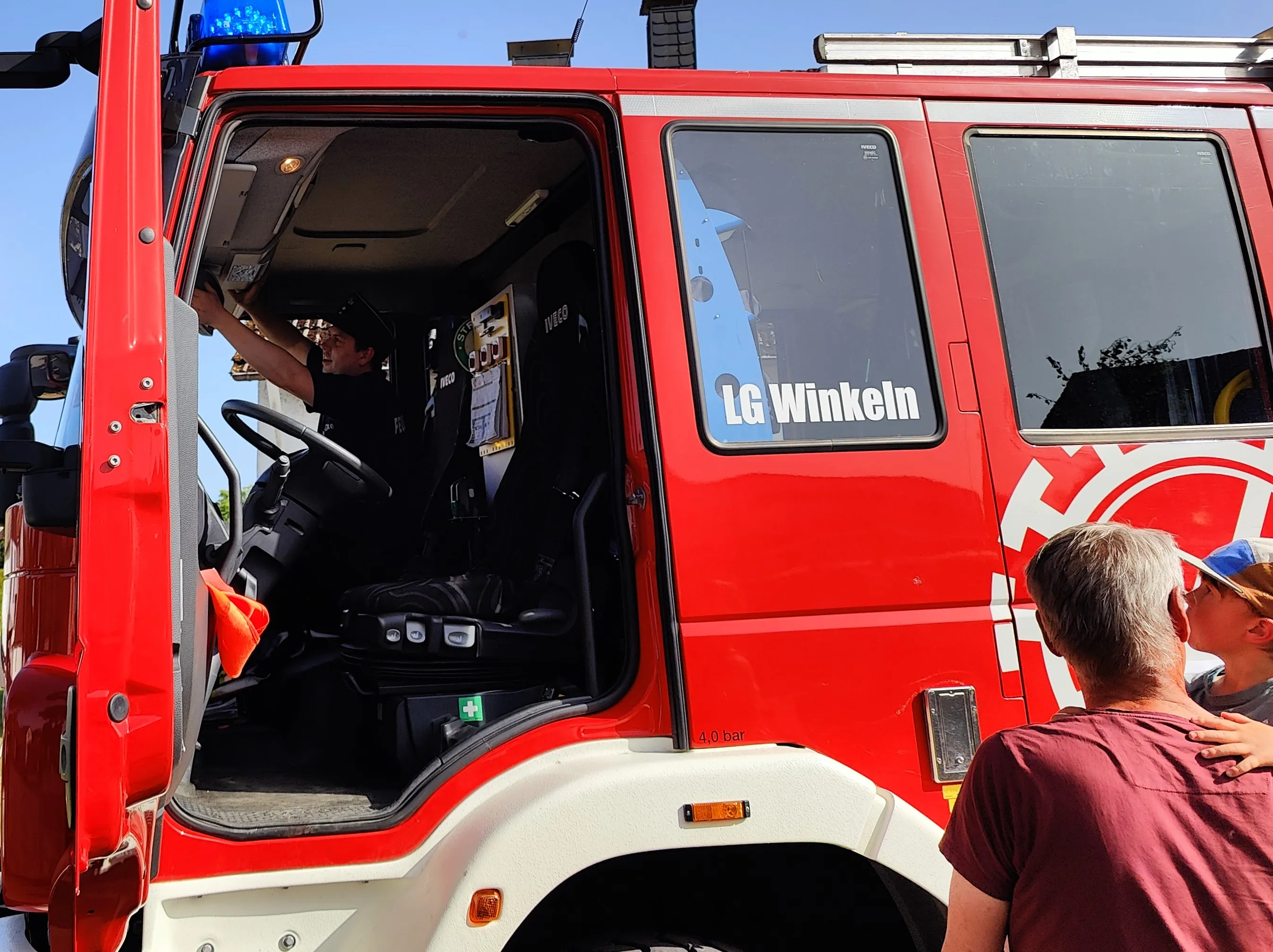 Ein Feuerwehrmann sitzt im Fahrerraum eines roten Feuerwehrautos, während ein Besucher mit grauem Haar und blauem Cap von hinten zu sehen ist. Auf der Tür steht „LG Winkeln“.