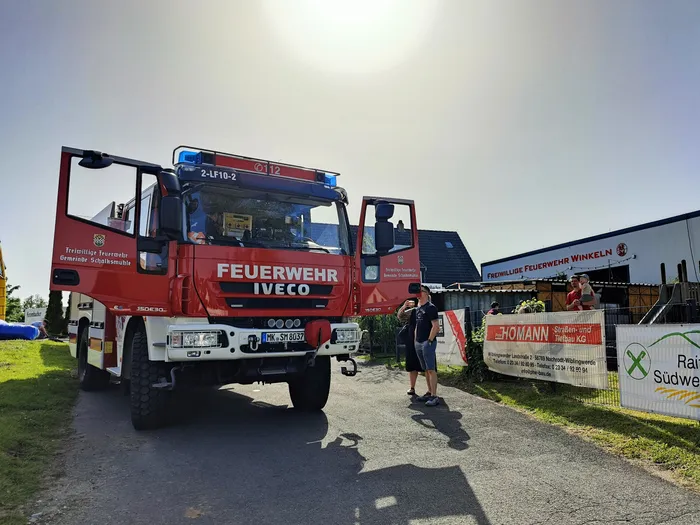 Roter Feuerwehrauto der Feuerwehr Schalksmühle mit der Aufschrift „FEUERWEHR IVECO“ steht auf einer Straße, während Menschen im Hintergrund stehen und ein Banner betrachten.