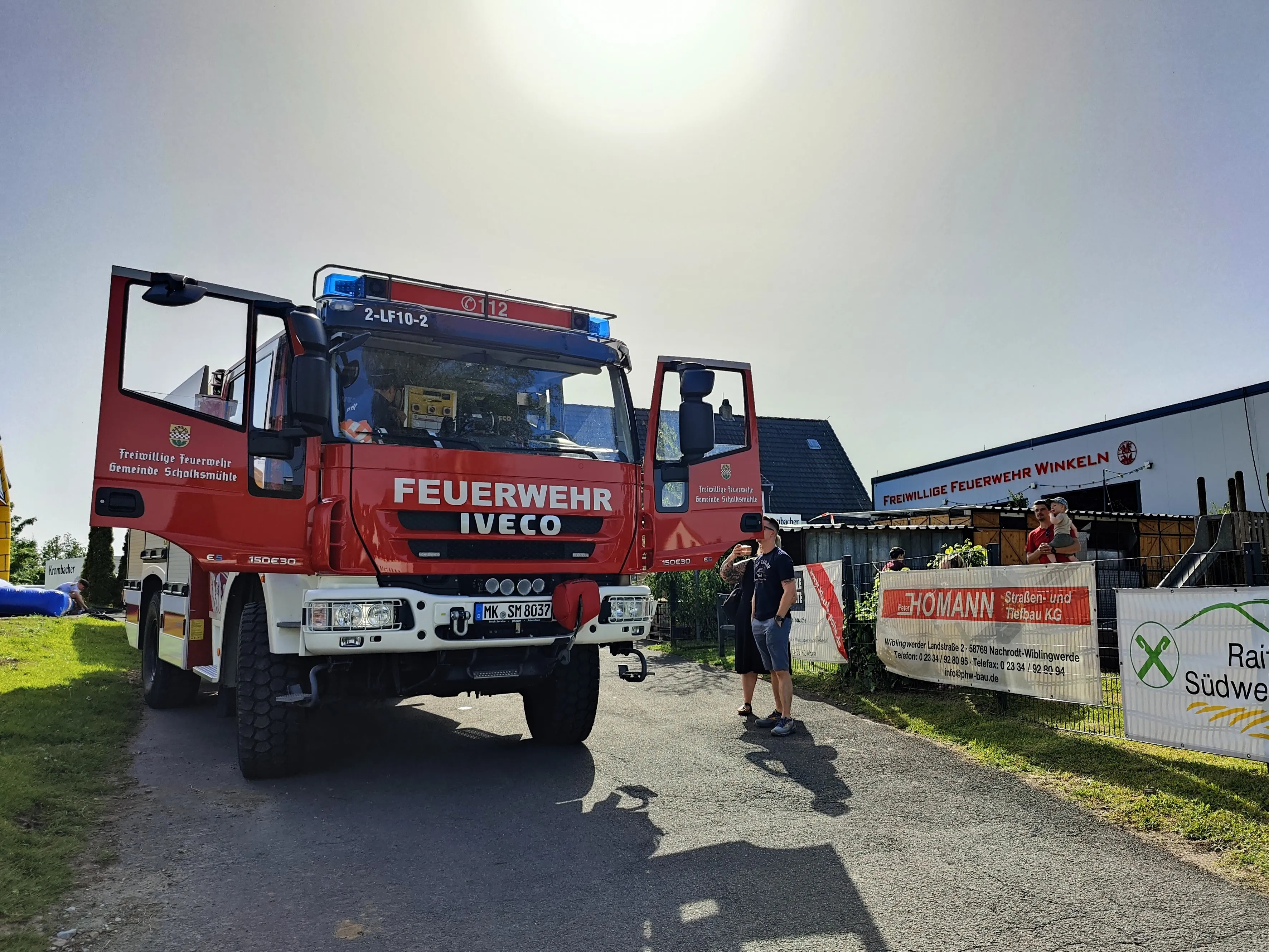 Roter Feuerwehrauto der Feuerwehr Schalksmühle mit der Aufschrift „FEUERWEHR IVECO“ steht auf einer Straße, während Menschen im Hintergrund stehen und ein Banner betrachten.