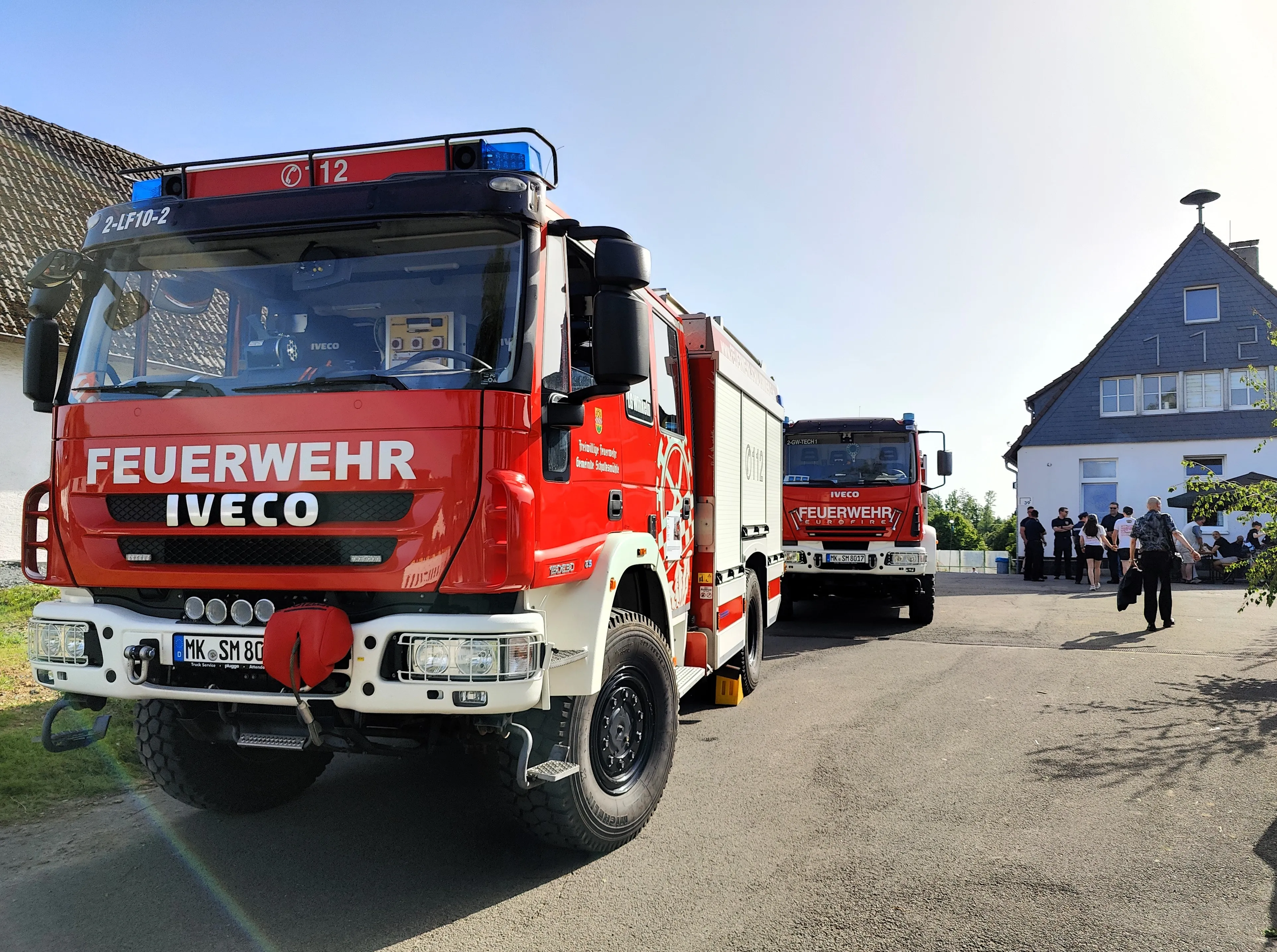 Zwei rote Feuerwehrautos der Feuerwehr Schalksmühle stehen auf einer Straße vor Häusern, im Hintergrund Menschen beim Dorffest.