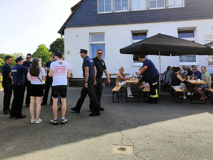 Menschengruppen vor einem Gebäude mit Biergartenbestuhlung, einige tragen Kleidung mit Aufdrucken und Uniformen der Feuerwehr.