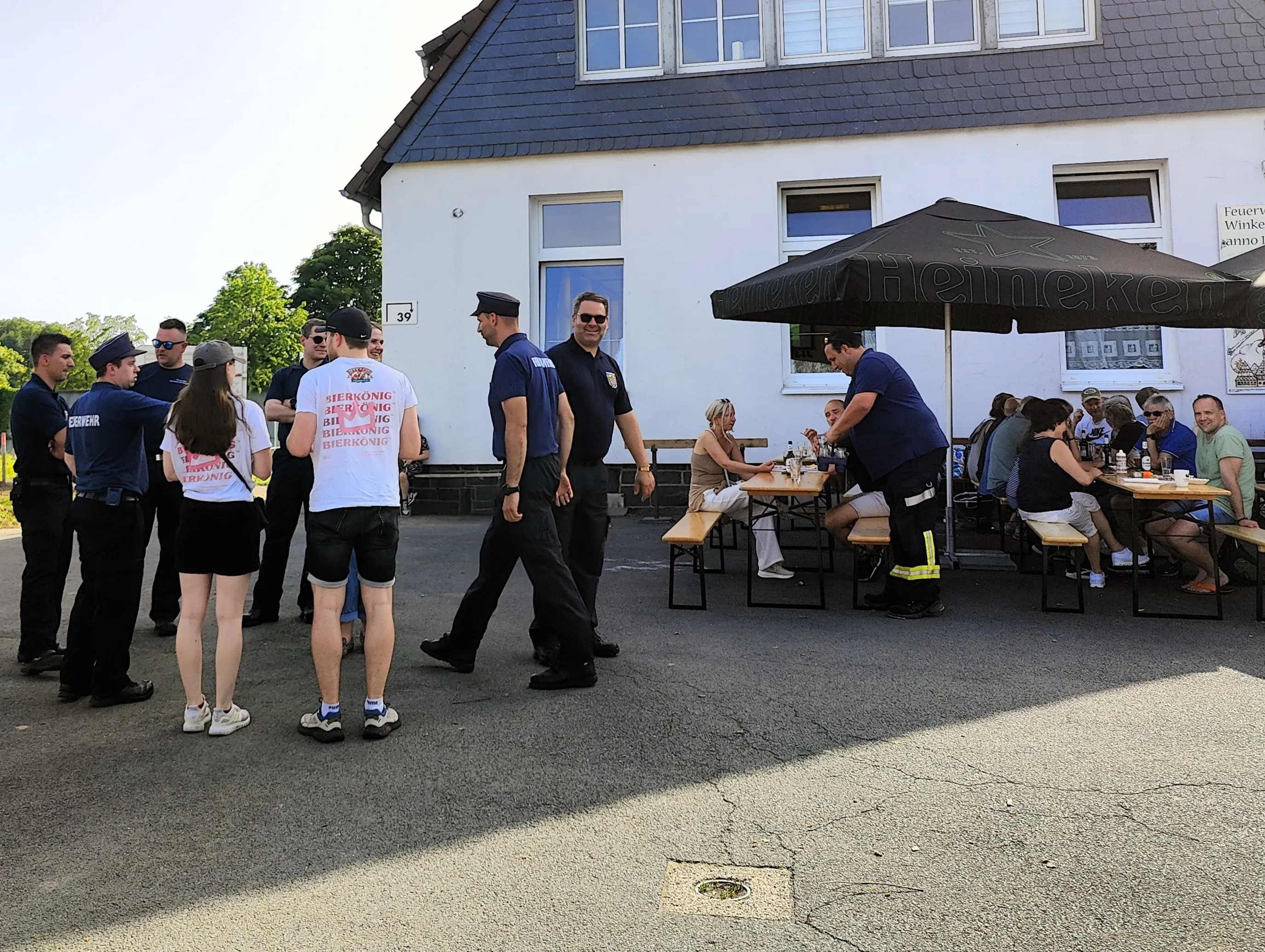 Menschengruppen vor einem Gebäude mit Biergartenbestuhlung, einige tragen Kleidung mit Aufdrucken und Uniformen der Feuerwehr.
