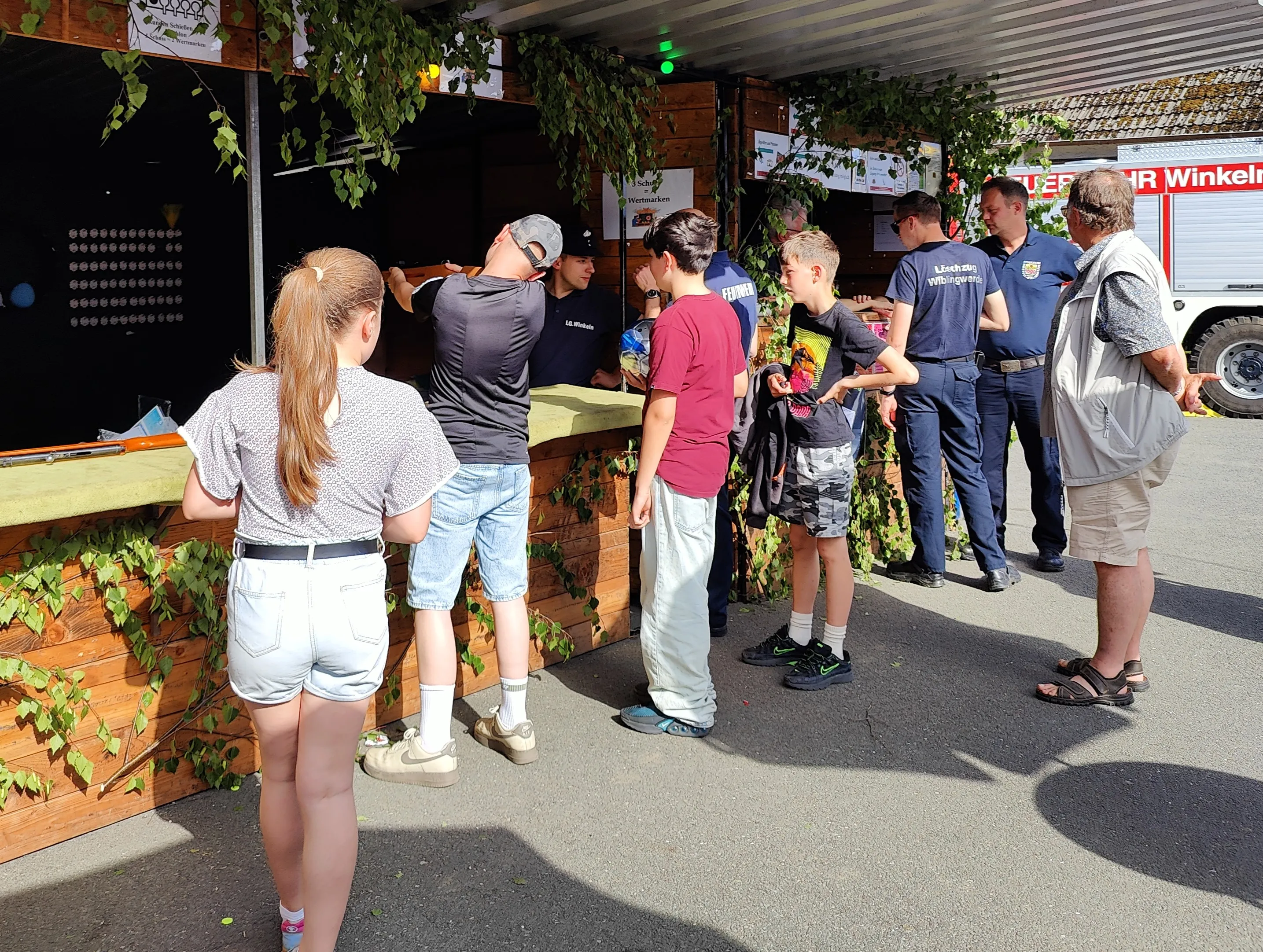 Eine Gruppe von Menschen, darunter Kinder und Feuerwehrleute, steht vor einem Holzstand auf einem Dorffest.