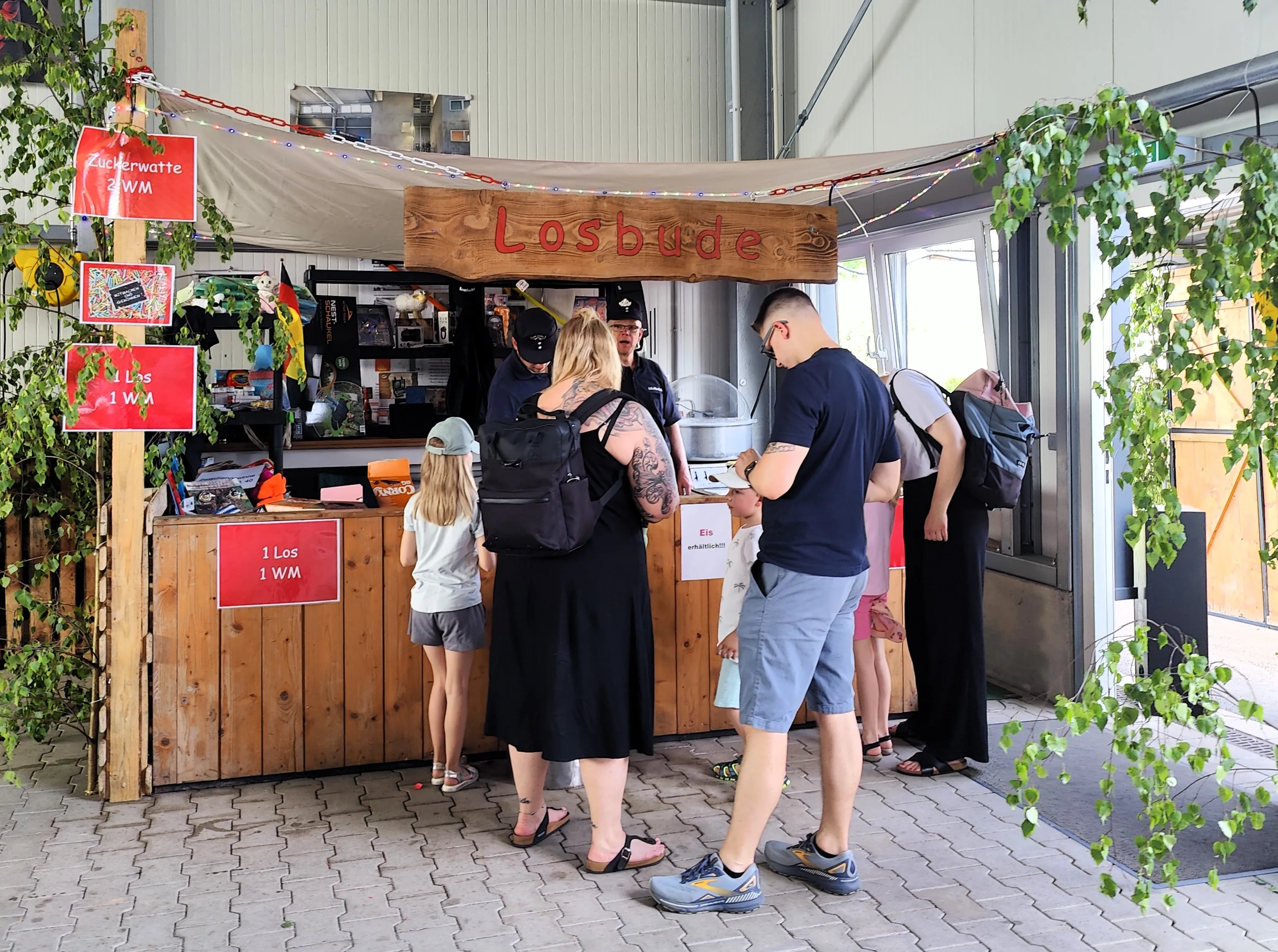 Menschen kaufen an einem Stand mit Süßigkeiten und Losen auf einem Dorffest. Über dem Stand hängt ein Schild mit der Aufschrift „Losbude“.