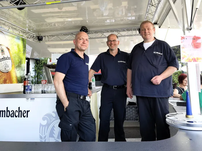 Drei Männer in dunkelblauen Poloshirts und Hosen stehen lächelnd vor einer Bar mit dem Schriftzug „Rombacher“.