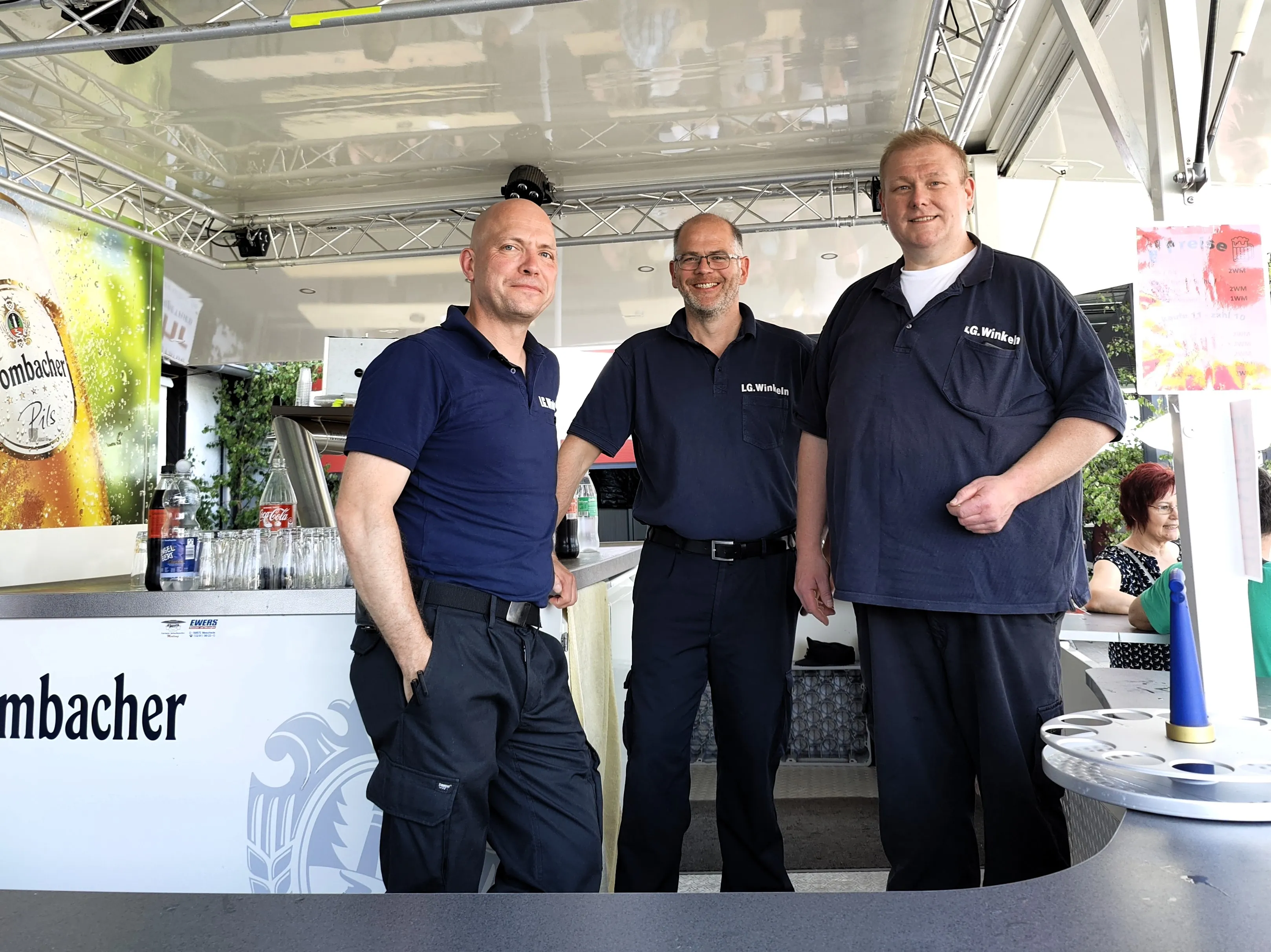 Drei Männer in dunkelblauen Poloshirts und Hosen stehen lächelnd vor einer Bar mit dem Schriftzug „Rombacher“.