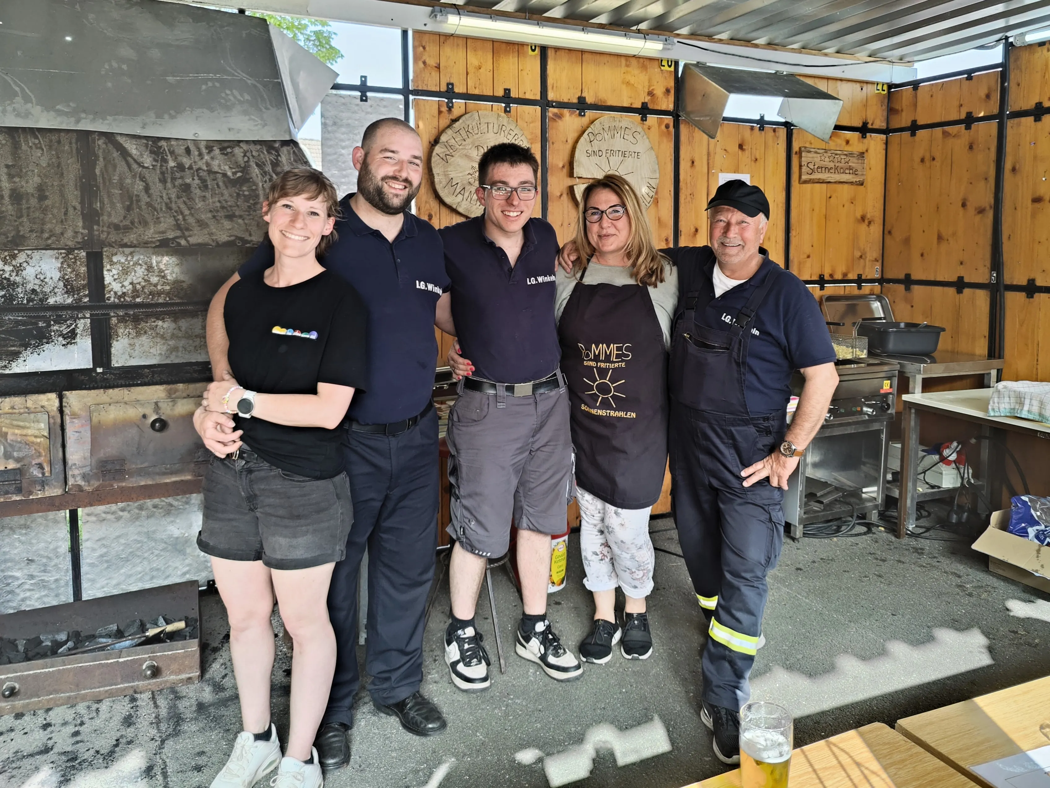 Fünf Personen stehen vor einem Holzschuppen mit der Aufschrift „Pommes“. Sie tragen Arbeitskleidung und lächeln in die Kamera.