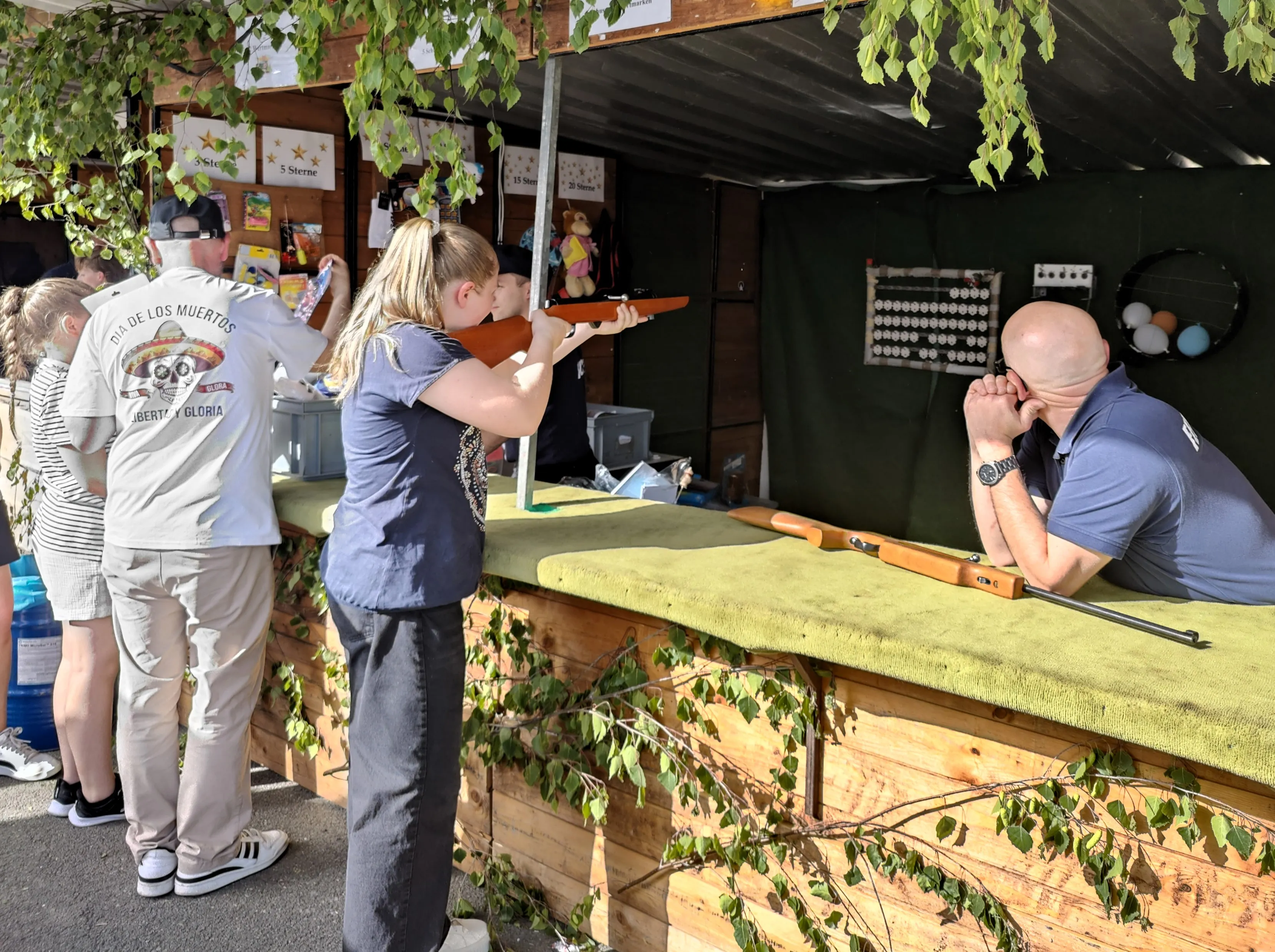 Zwei Personen zielen mit Gewehren auf eine Zielscheibe auf einem Schießstand während eines Dorffestes. Im Hintergrund stehen weitere Besucher und eine Reihe von Stofftieren.