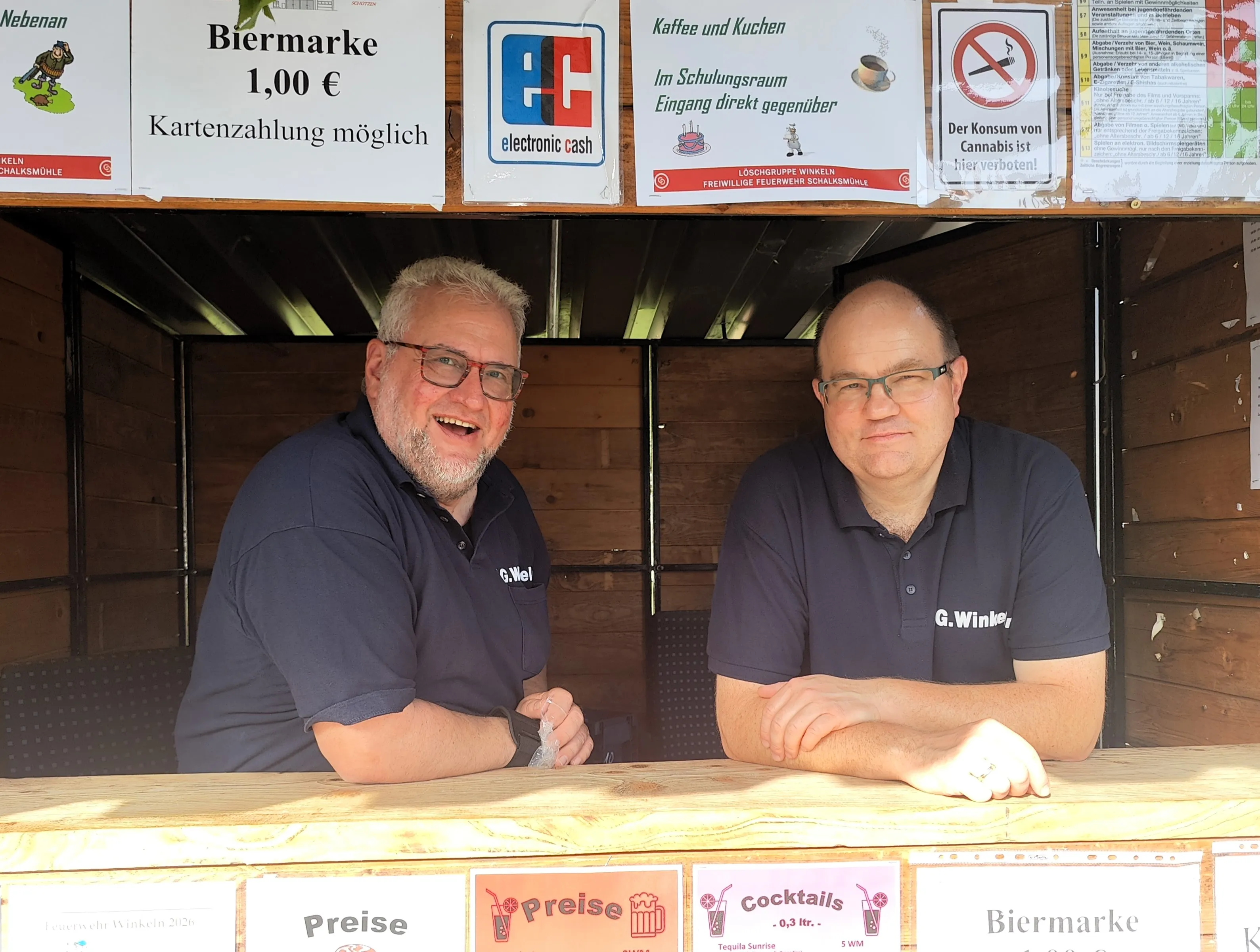 Zwei Männer sitzen hinter einem Tresen in einem hölzernen Stand, lächeln in die Kamera. Im Hintergrund hängen Schilder mit Preisen für Bier und Cocktails sowie Hinweise zum Dorffest.