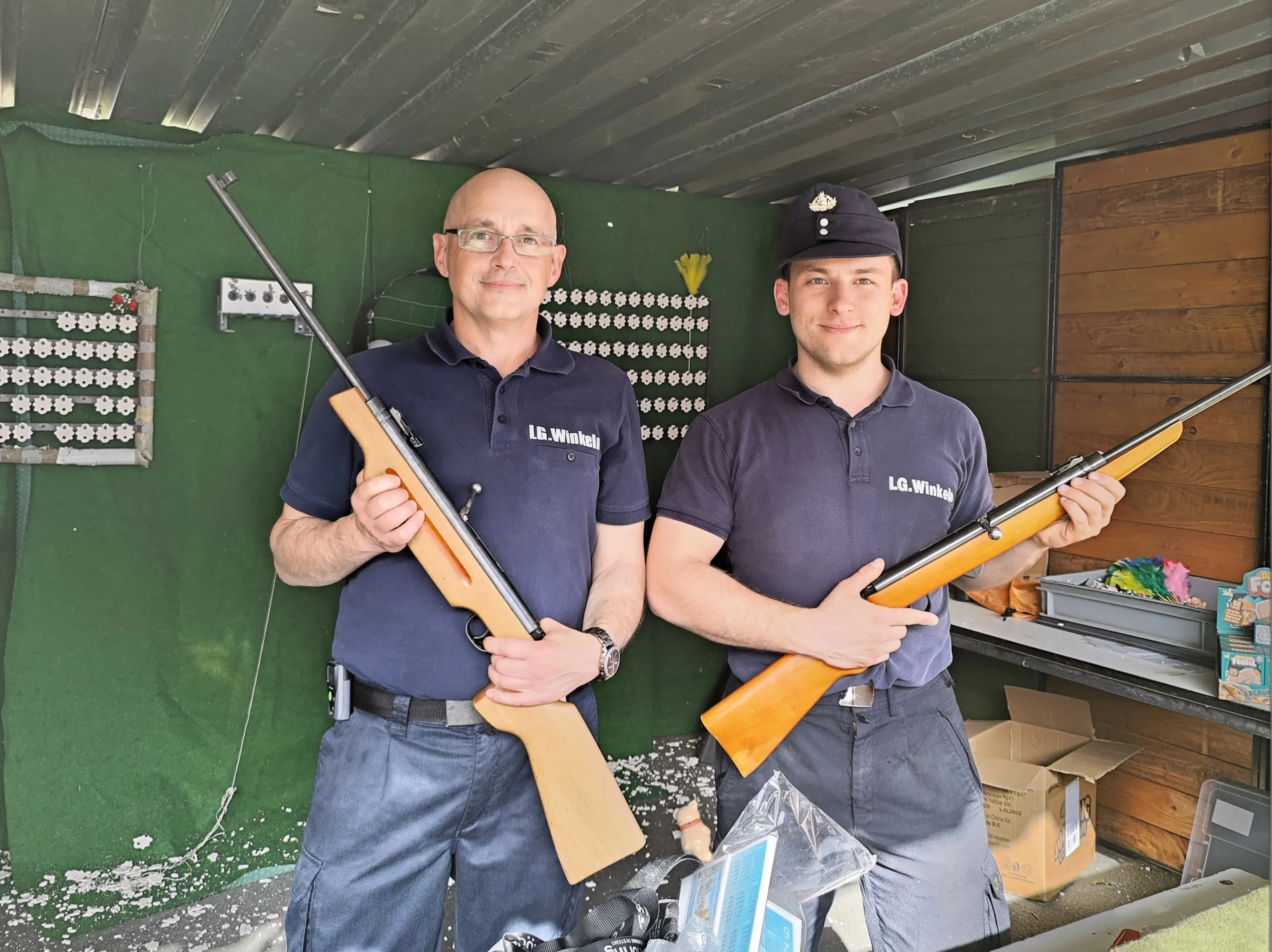 Zwei Männer in dunkelblauen Uniformen mit dem Schriftzug „LG.Winkel“ halten jeweils ein Gewehr. Im Hintergrund eine grüne Wand mit mehreren Vogelkäfigen.