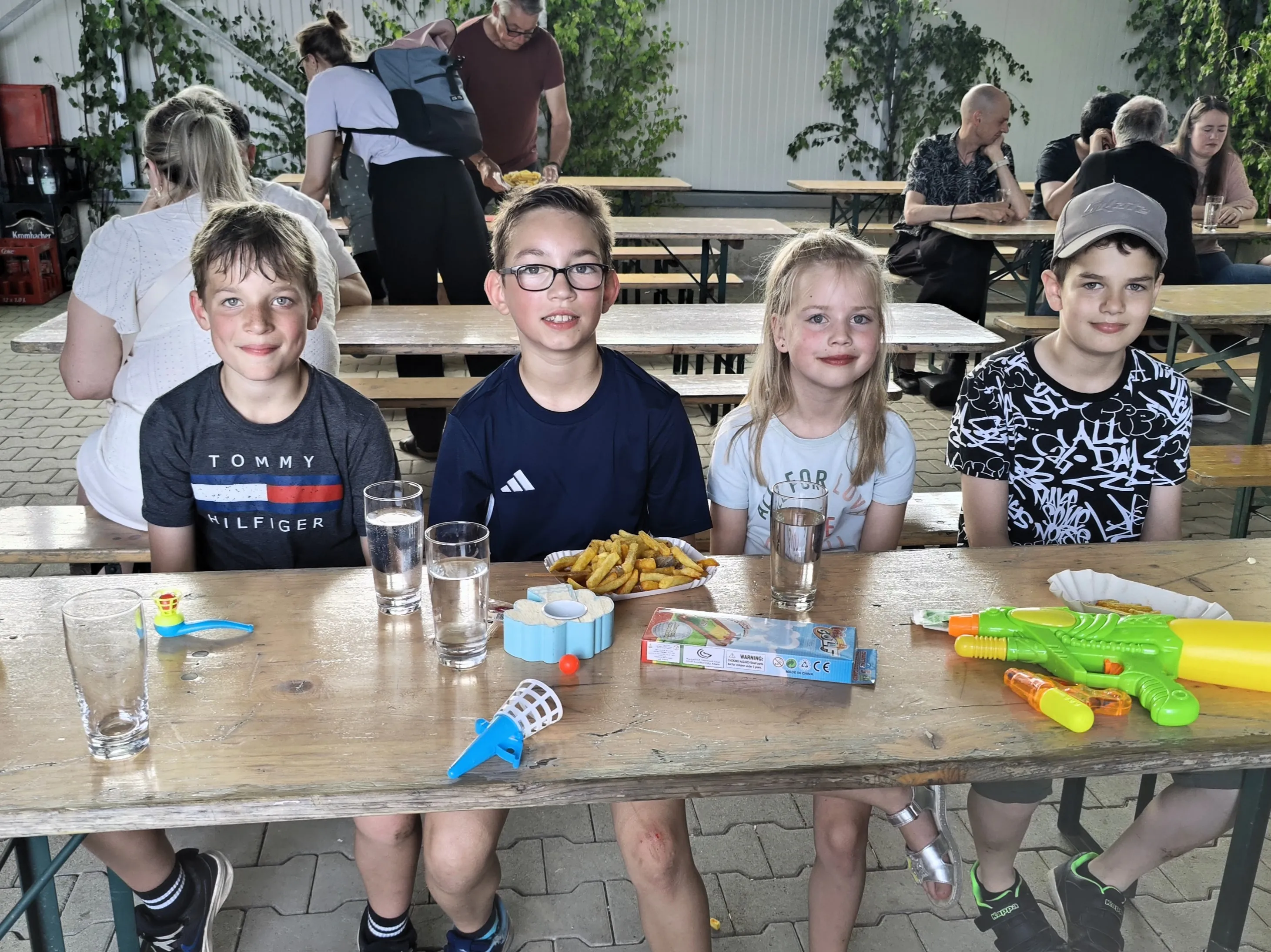 Vier Kinder sitzen an einem Holztisch mit Pommes und Getränken, umgeben von Spielzeug. Im Hintergrund sind weitere Besucher eines Dorffestes zu sehen.