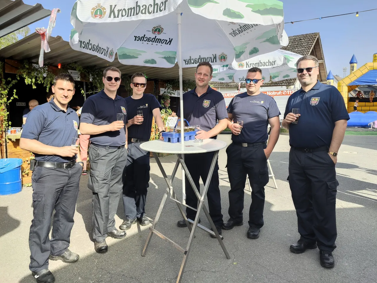 Sechs Feuerwehrleute in Uniform stehen lächelnd um einen runden Stehtisch mit Getränken auf einem Dorffest unter Sonnenschirmen mit Werbeaufdruck.