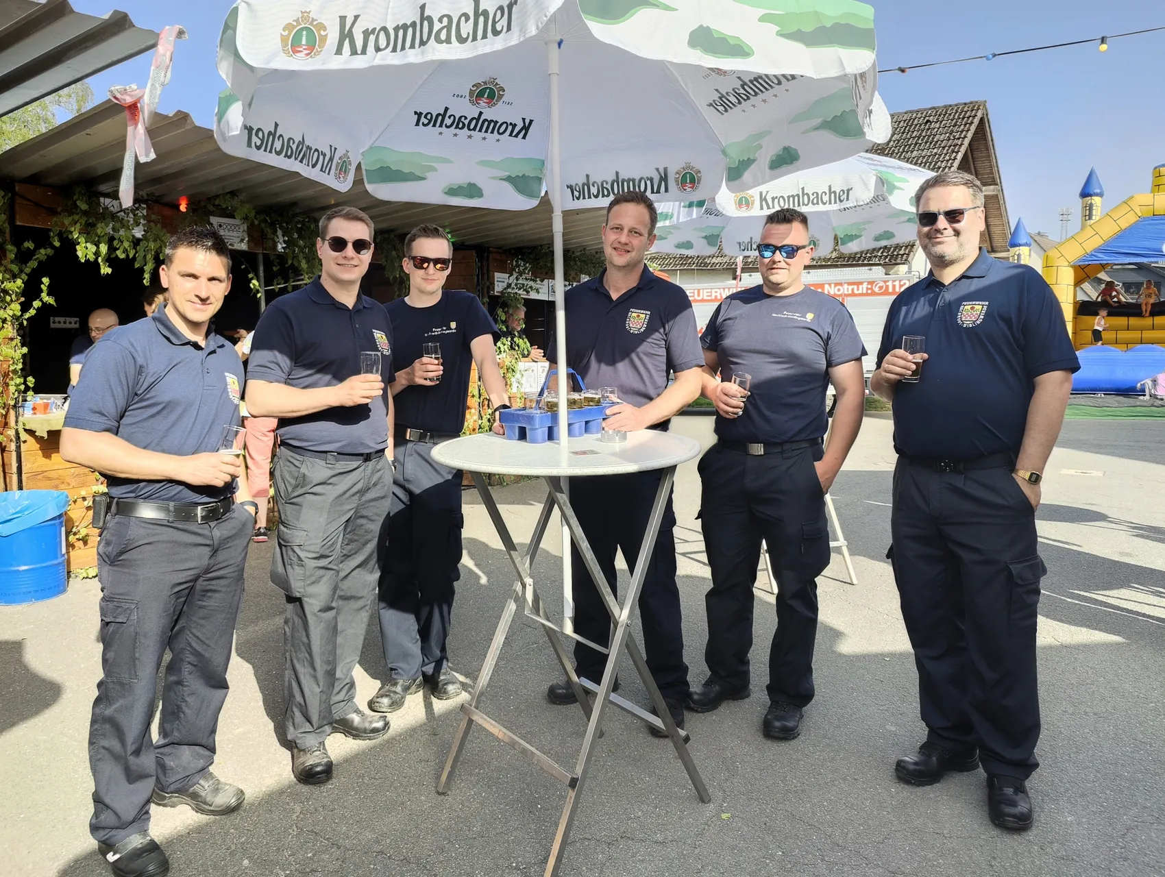Sechs Feuerwehrleute in Uniform stehen lächelnd um einen runden Stehtisch mit Getränken auf einem Dorffest unter Sonnenschirmen mit Werbeaufdruck.
