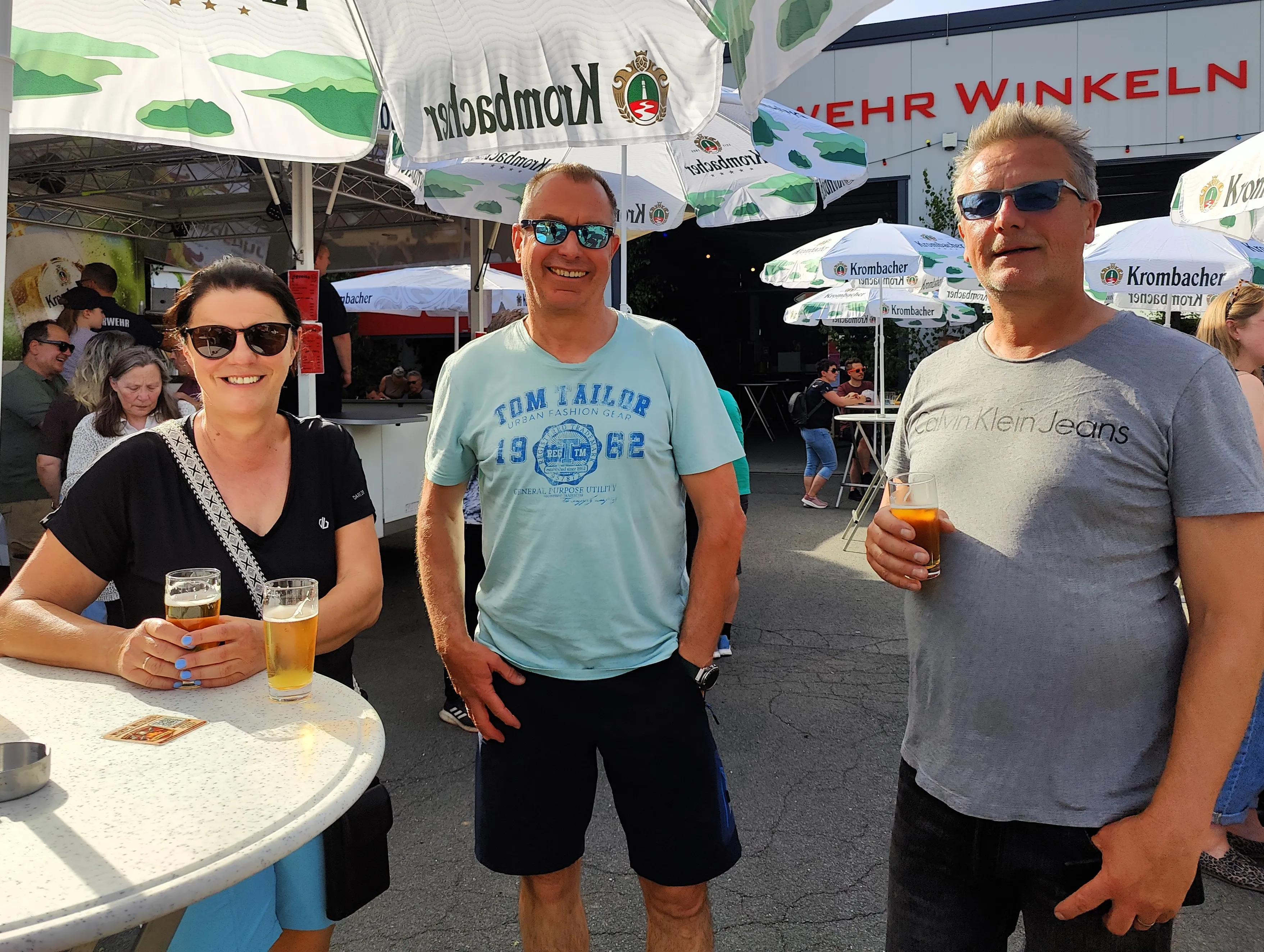Drei Personen stehen lächelnd vor Sonnenschirmen mit dem Krombacher-Logo und halten Getränke in der Hand. Im Hintergrund sind weitere Besucher eines Dorffestes zu sehen.