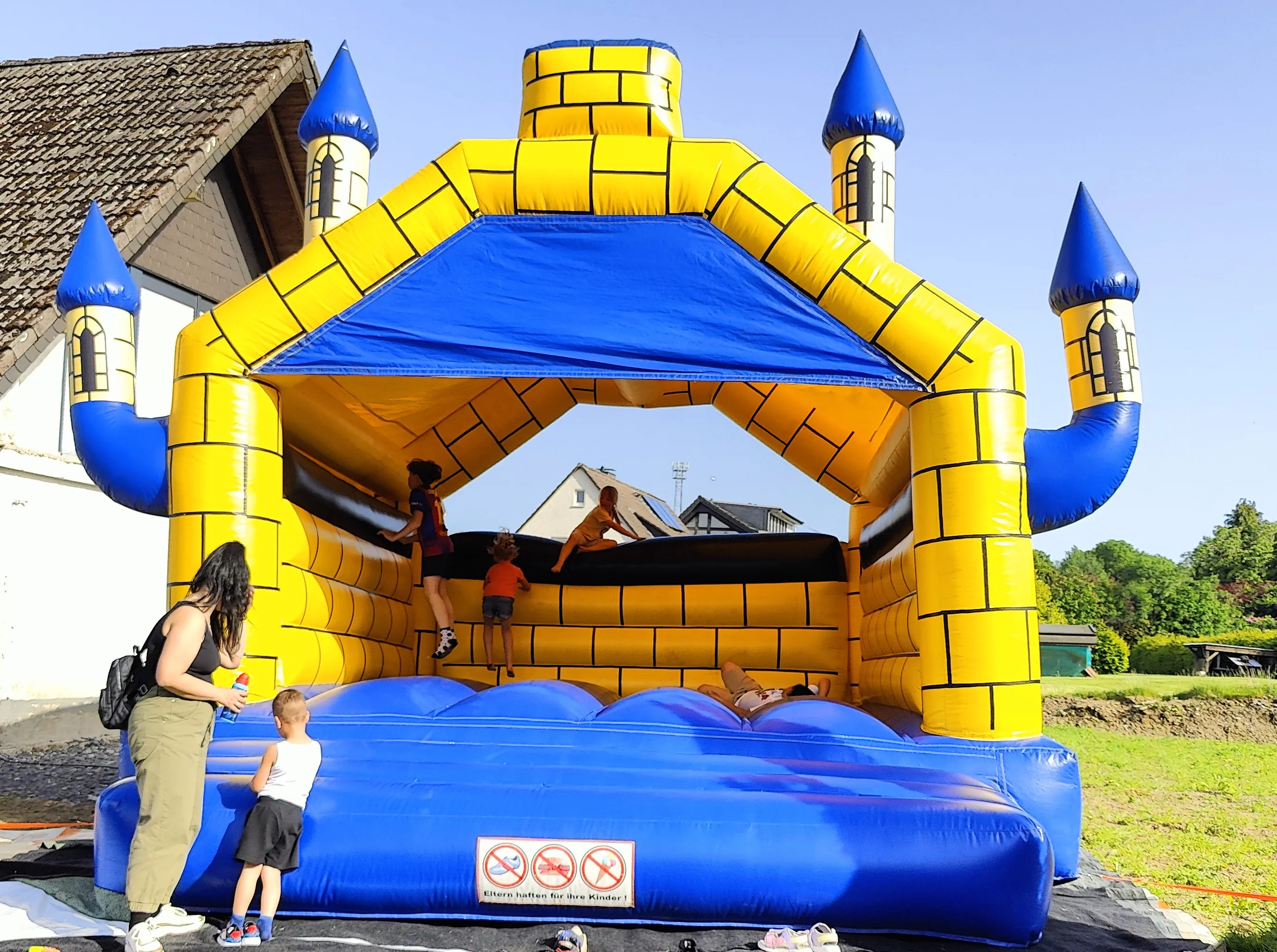 Eine große, blaue und gelbe Hüpfburg in Form eines Schlosses steht auf einer Wiese. Kinder spielen und klettern auf der Burg, während eine Frau am Rand steht und zusieht.