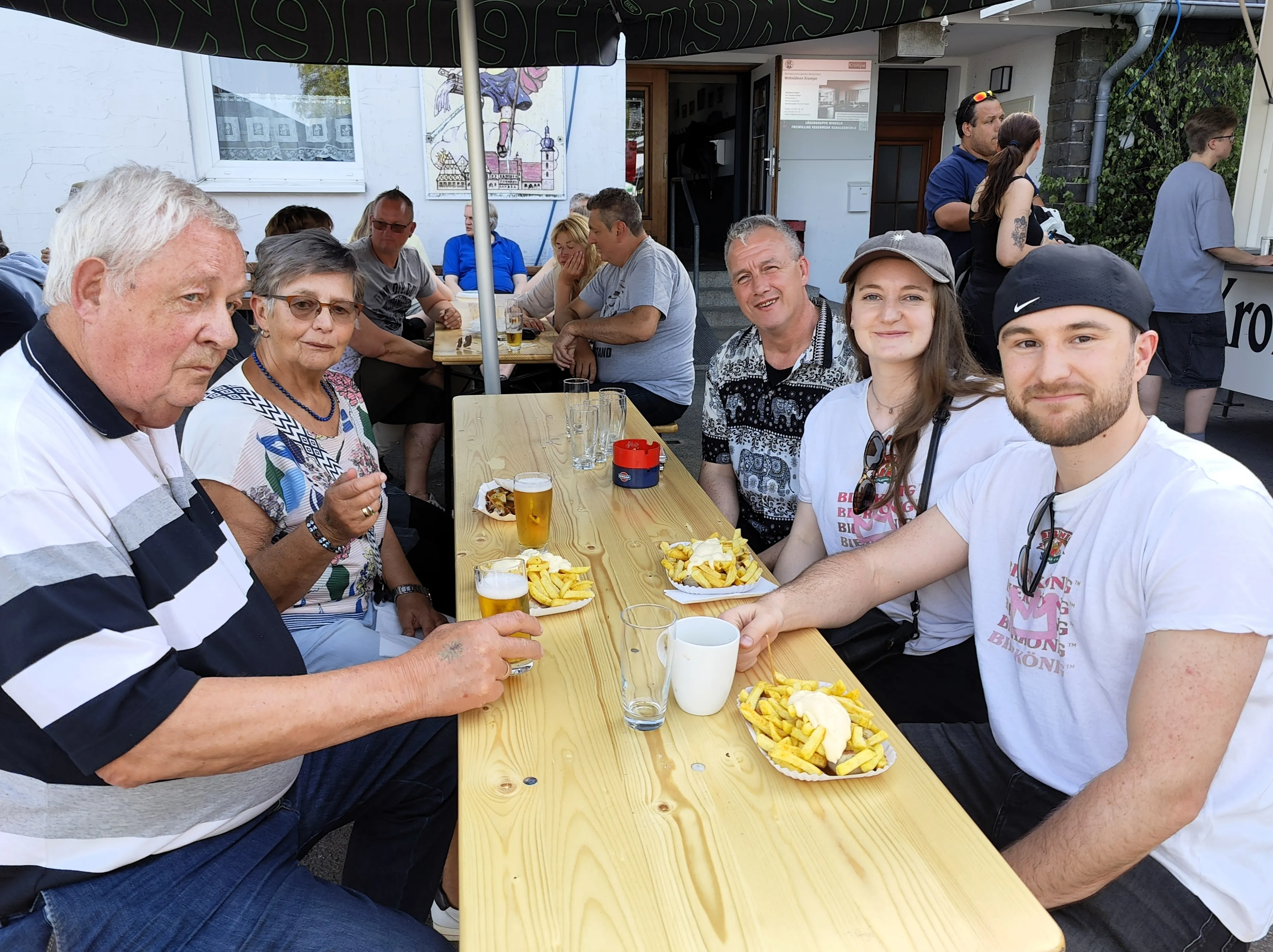 Mehrere Personen sitzen an einem Holztisch im Freien, essen Pommes und trinken Bier. Im Hintergrund sind weitere Besucher eines Dorffestes zu sehen.
