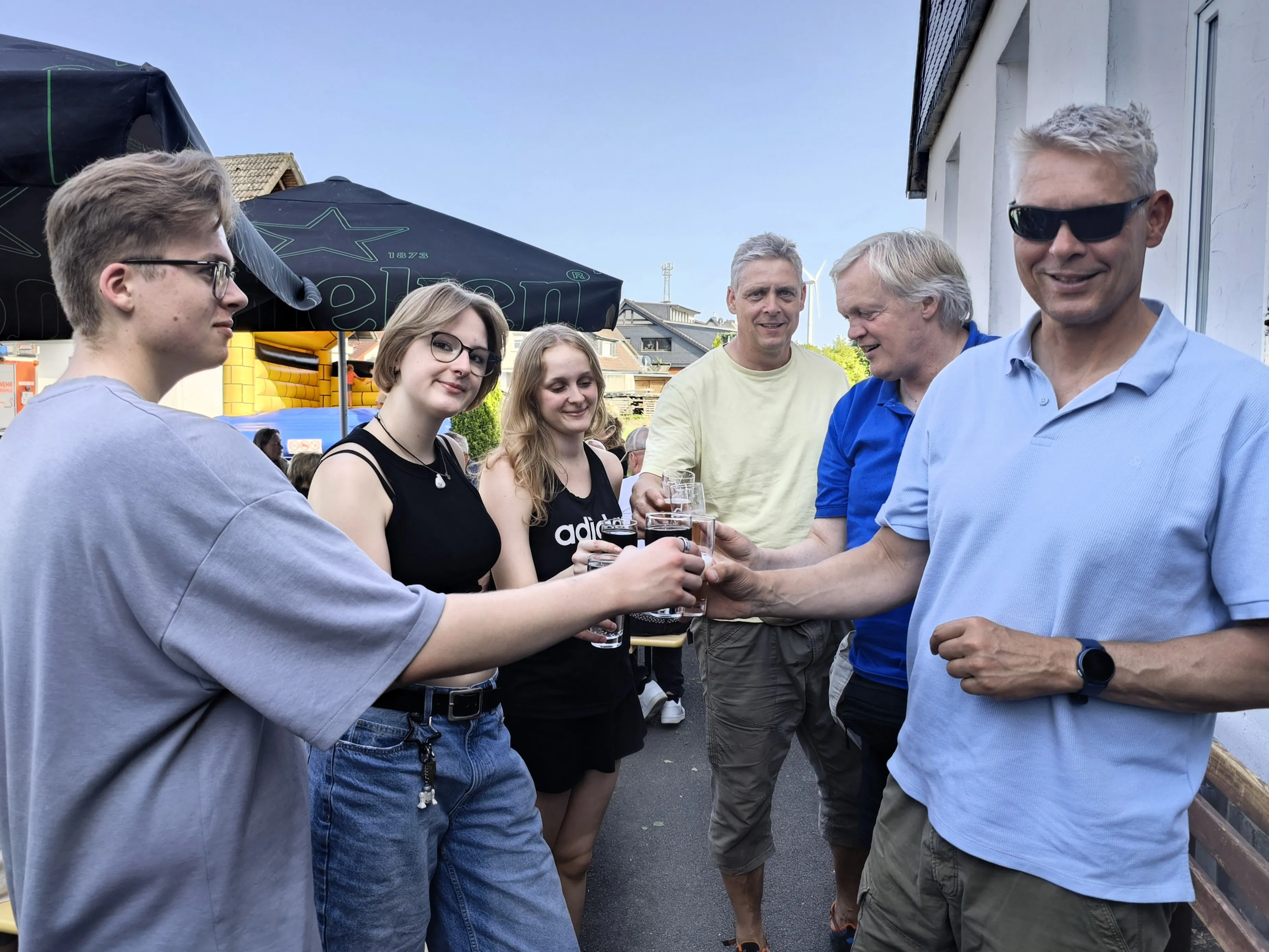 Eine Gruppe von sechs Personen steht im Freien und prostet sich mit Biergläsern zu. Im Hintergrund ein dunkler Pavillon und Häuserfassaden.