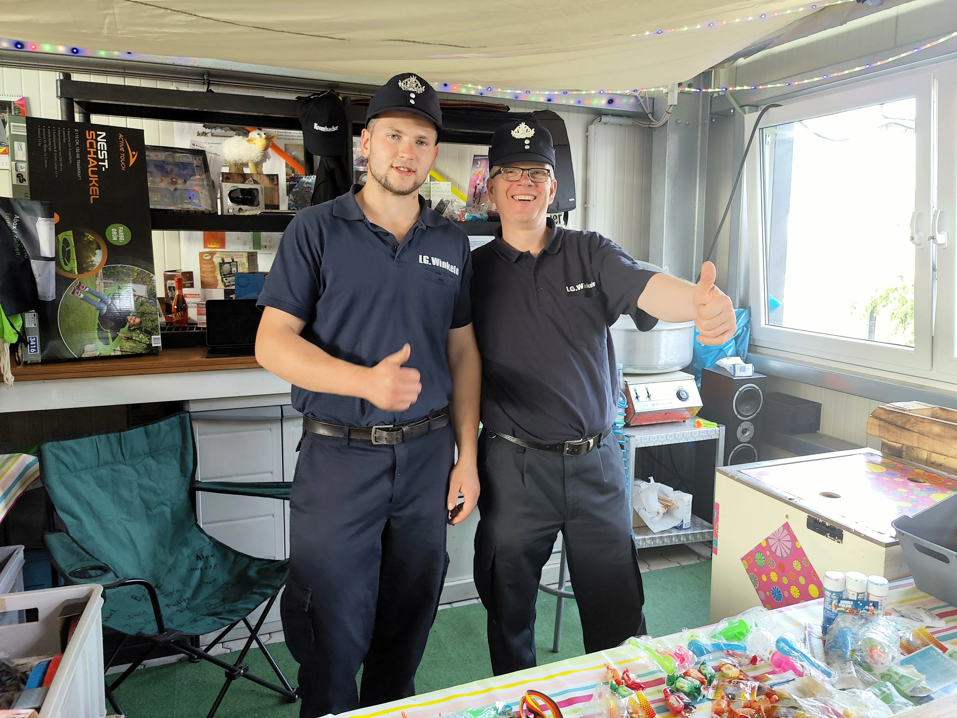 Zwei Feuerwehrleute in Uniformen mit Kappen und Namensschildern geben Daumen hoch. Im Hintergrund ein Stand mit Verkaufsartikeln und Dekorationen.