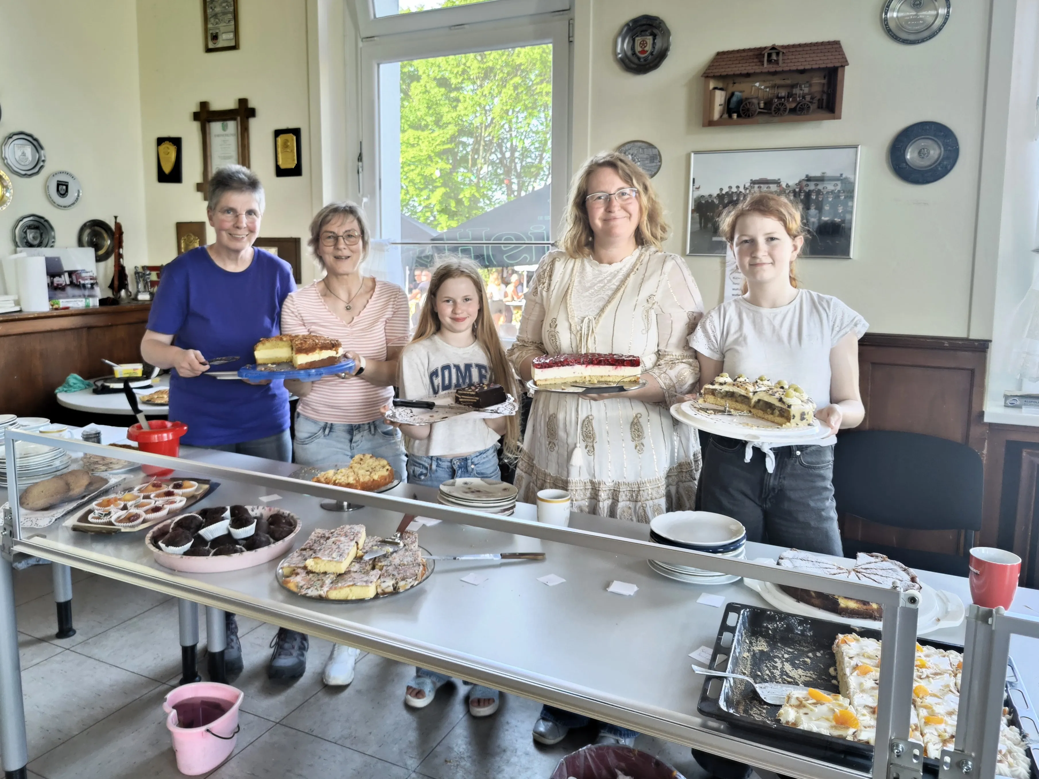 Fünf Personen stehen hinter einem Tisch mit einer großen Auswahl an Kuchen und Torten. Im Hintergrund hängen Bilder und Dekorationen an der Wand.