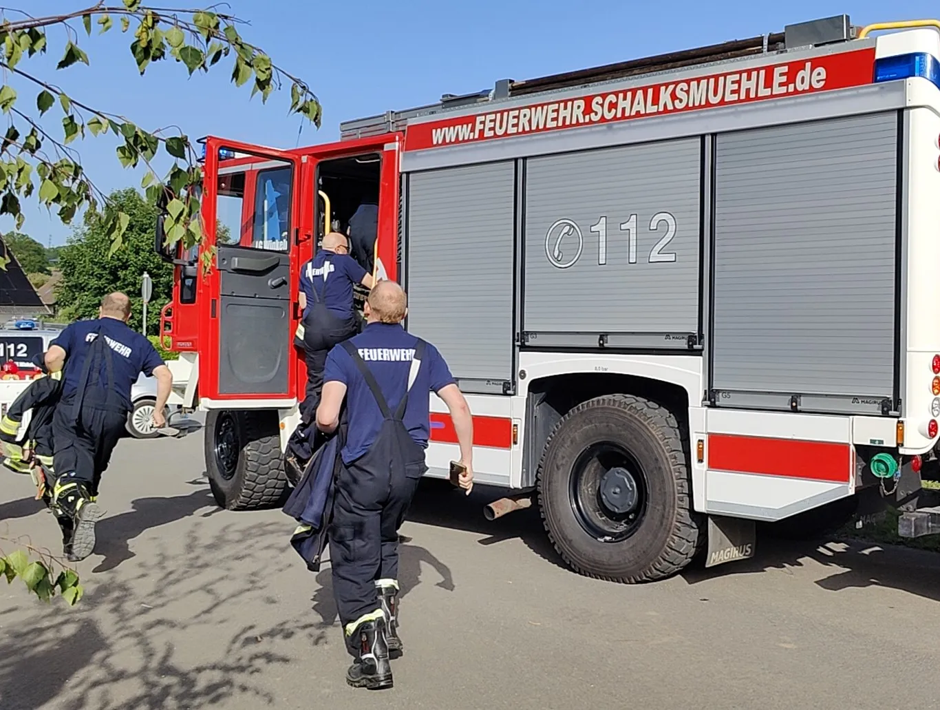 Feuerwehrleute steigen von einem roten Feuerwehrauto ab, während im Hintergrund ein Dorffest stattfindet.